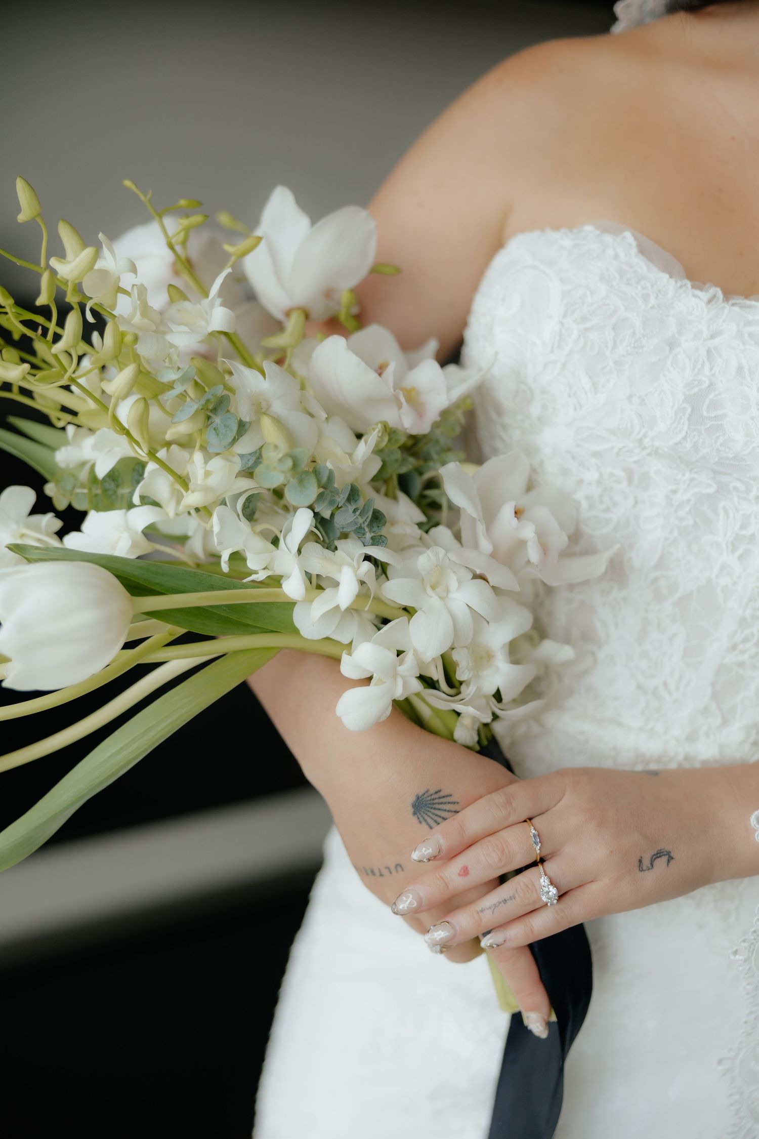 Bride holding elegant white bouquet of orchids and tulips with diamond engagement ring and delicate tattoos visible.