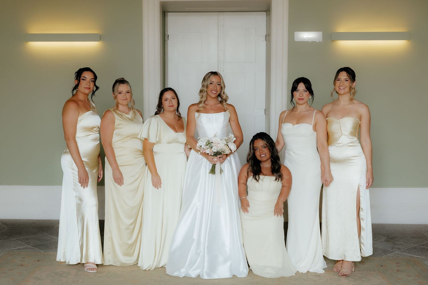A group of people wearing white and cream colored formal dresses pose together for a wedding party photo.