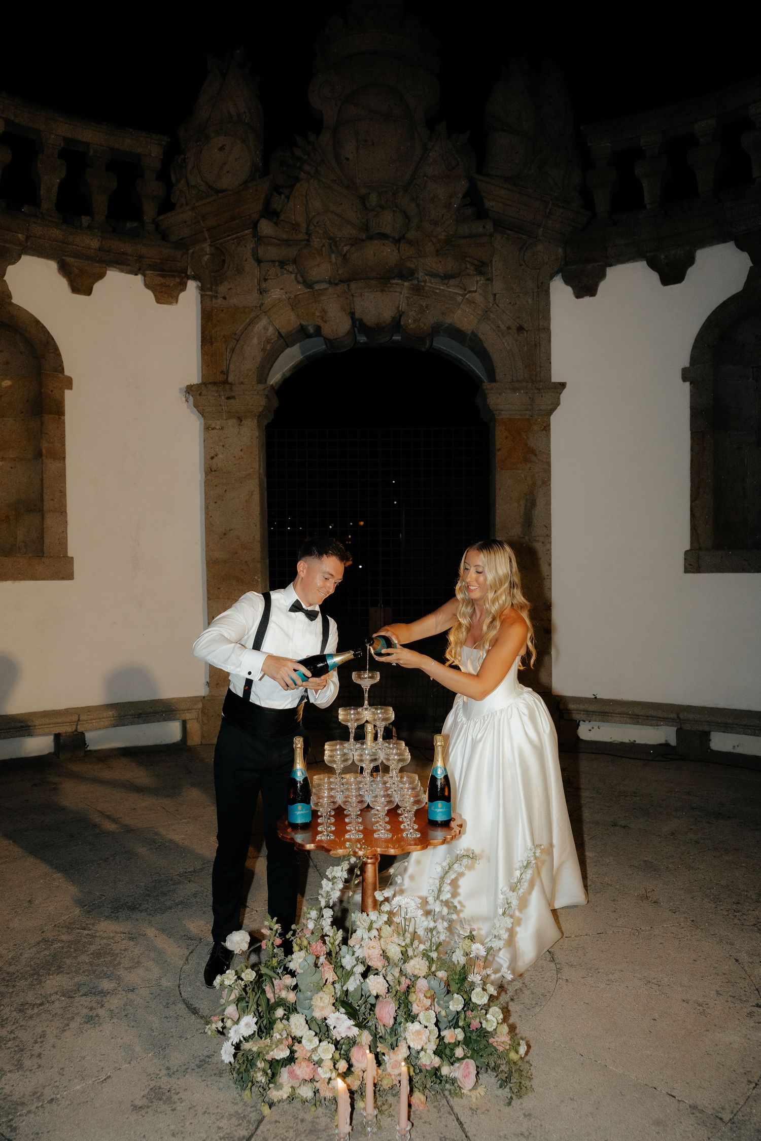 A couple pours champagne into a tower of glasses decorated with floral arrangements at an elegant venue.