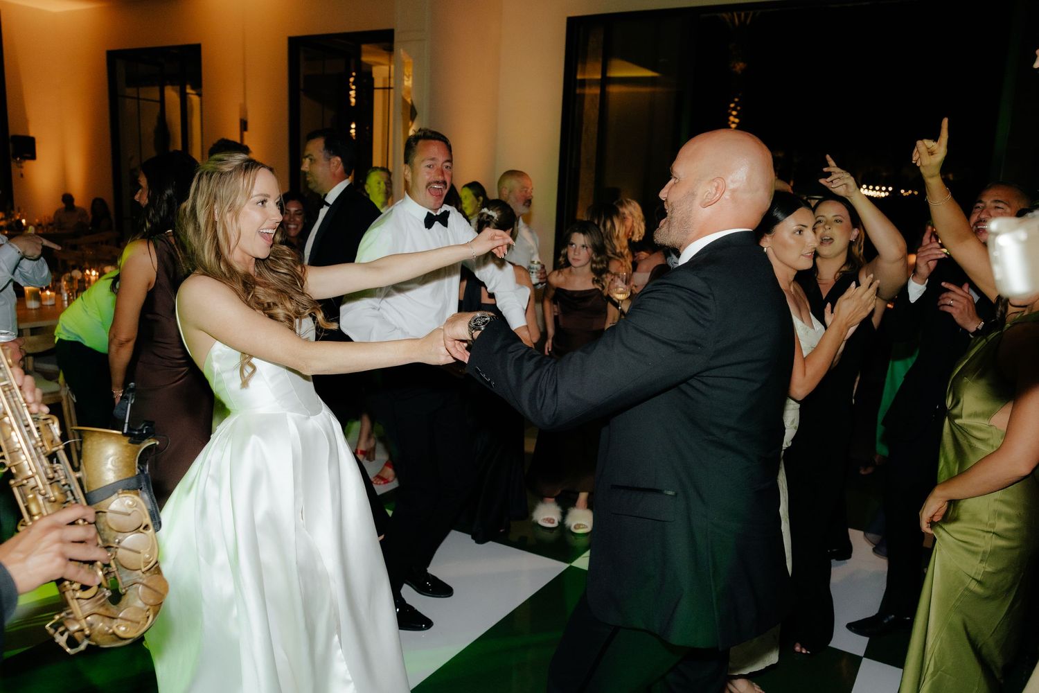 Bride and groom dance energetically on the dance floor with a live saxophonist and celebrating wedding guests nearby.