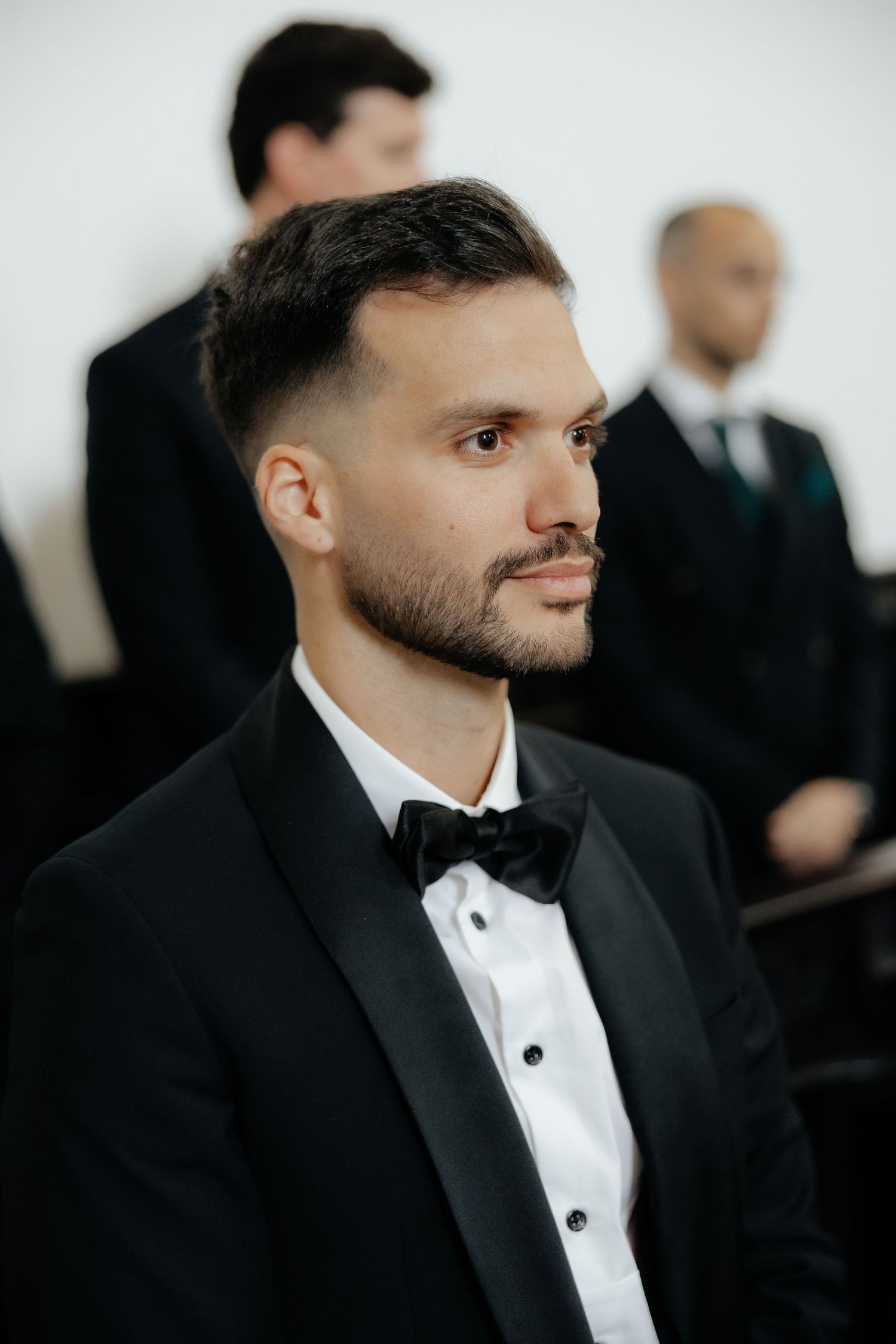 Handsome groom in black tuxedo with bow tie seated during wedding ceremony.