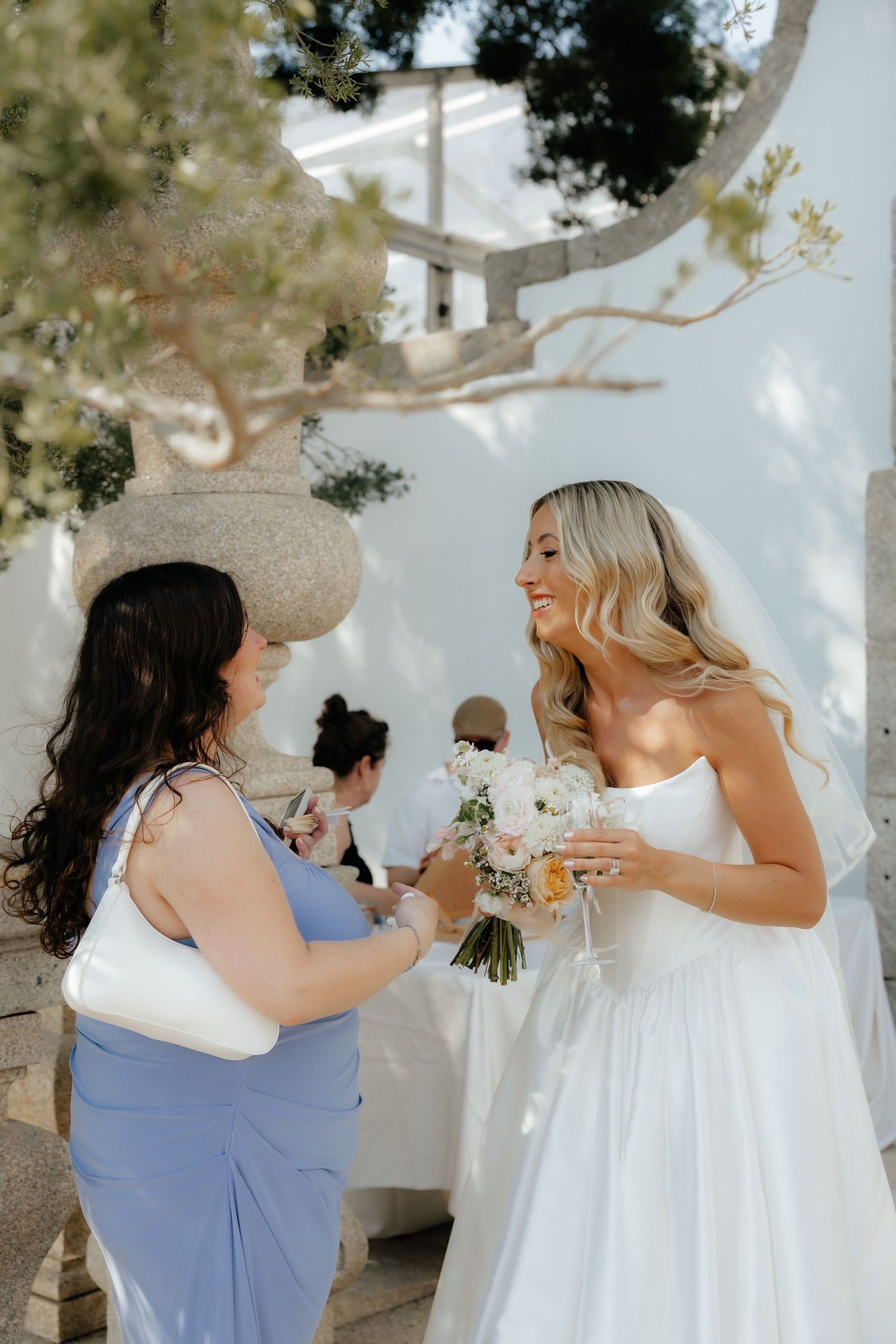Smiling bride in white strapless gown holds bouquet while chatting with bridesmaid in blue dress.
