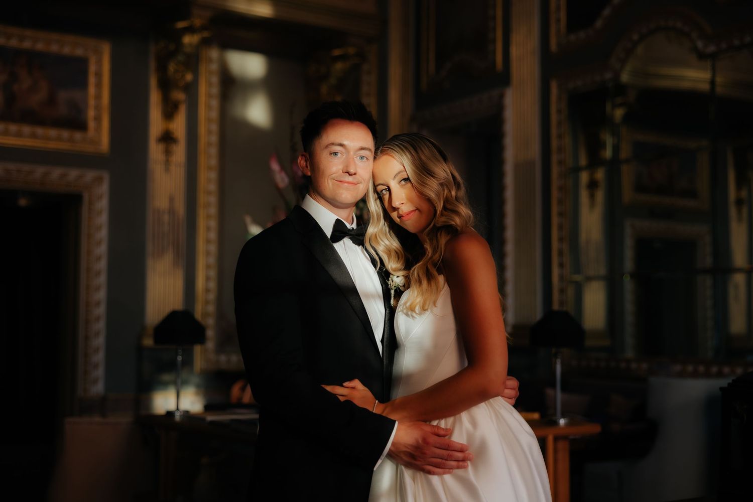 Romantic portrait of bride and groom embracing in warm golden light inside a grand ornate ballroom.