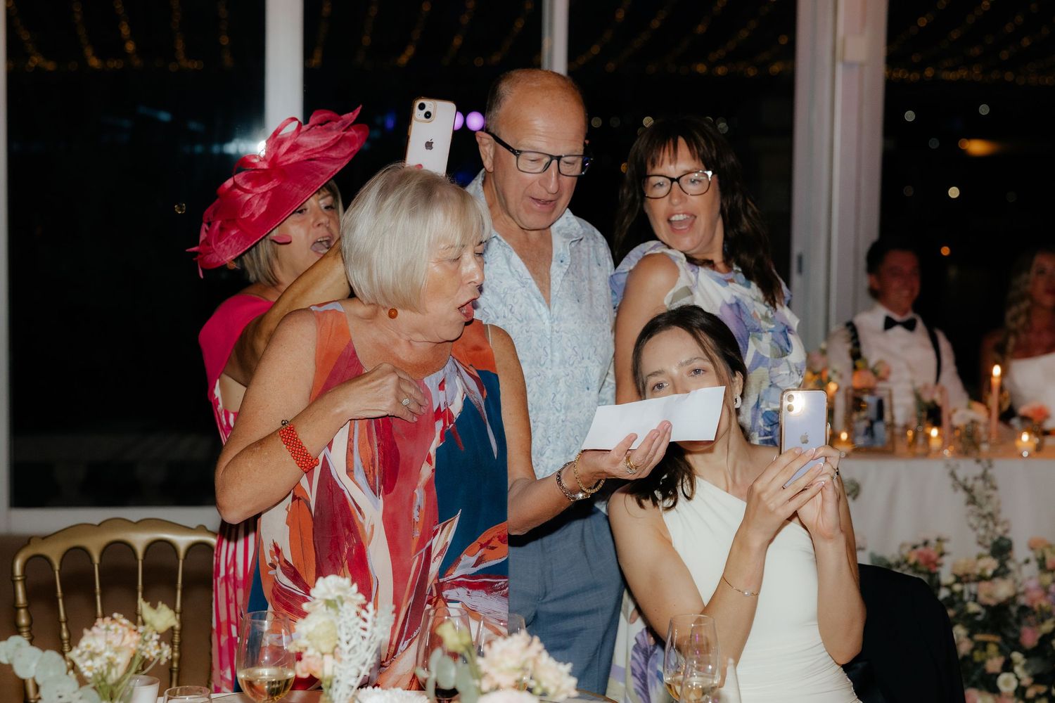 A group of wedding guests in formal attire share laughter and conversation at an elegant candlelit reception dinner.