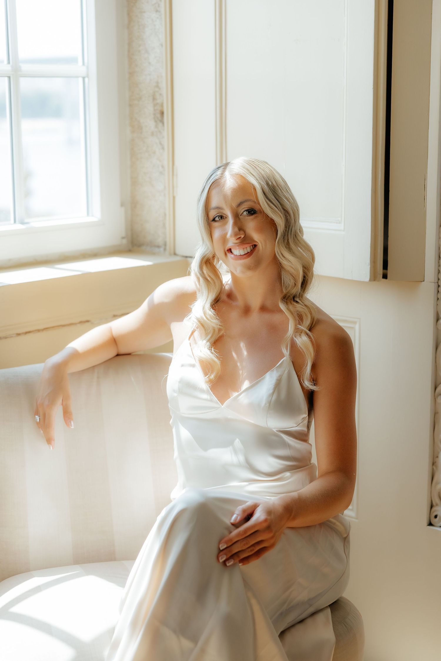 A person in a white silk dress poses gracefully by a sunlit window in an ethereal indoor photoshoot.