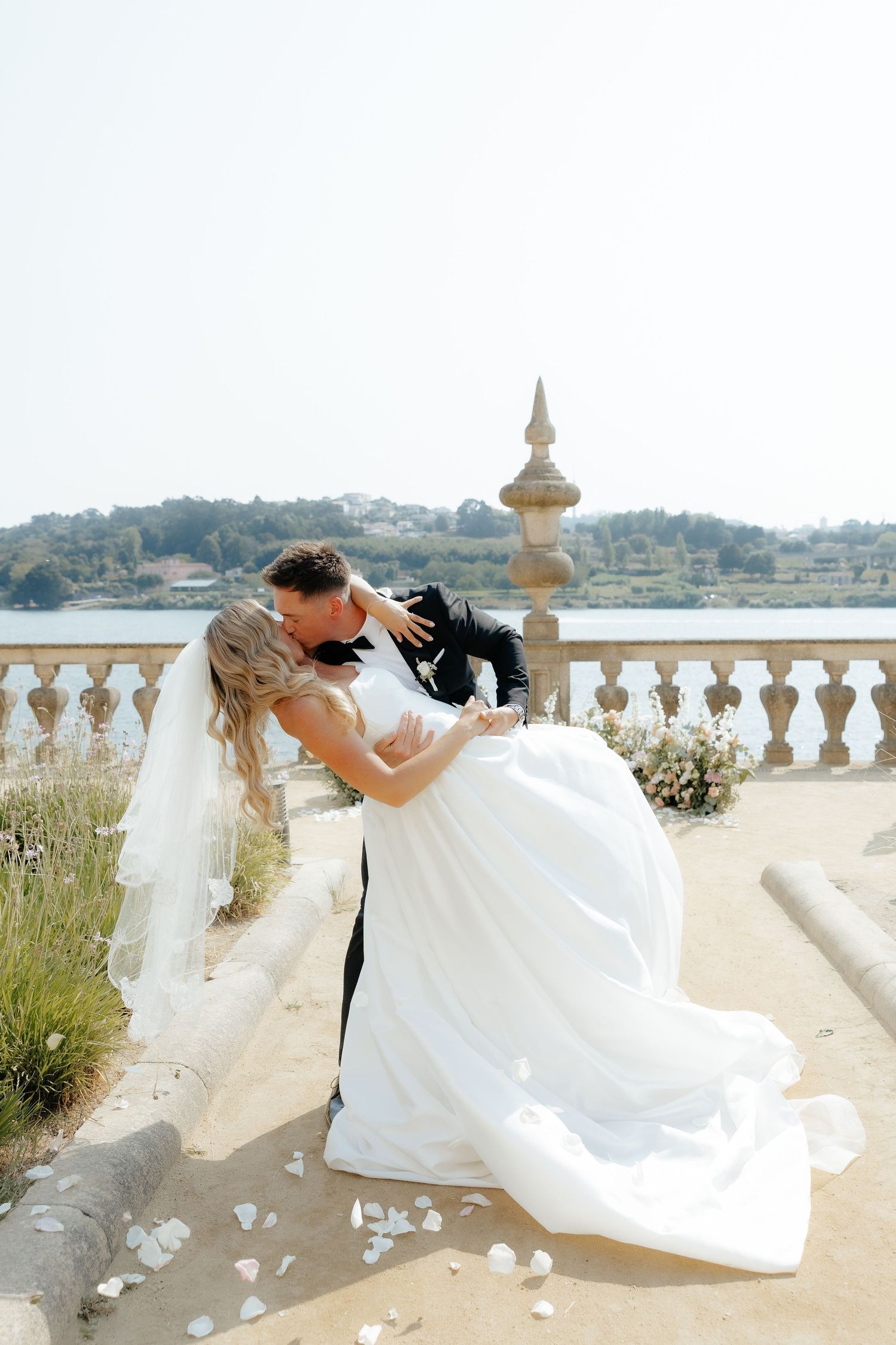 A romantic wedding couple dips in a dramatic pose along a stone balustrade with water views in the background.