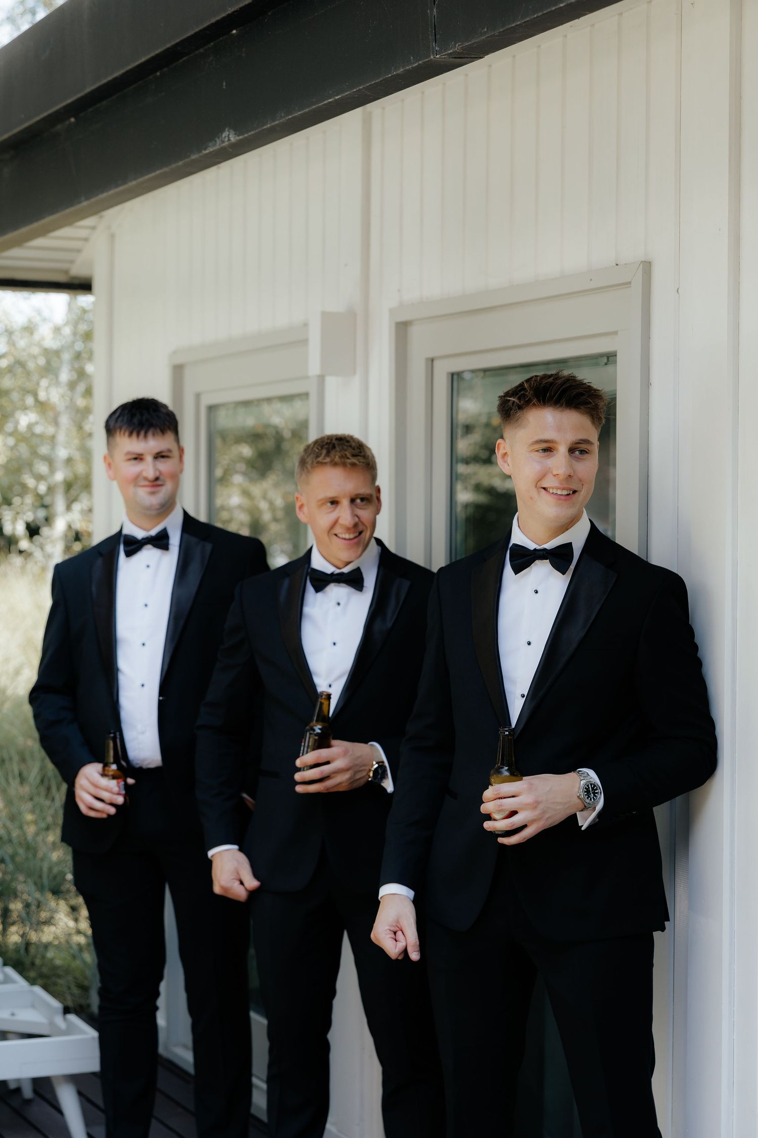 Groomsmen in black tuxedos and bow ties share laughs while holding drinks during wedding festivities.