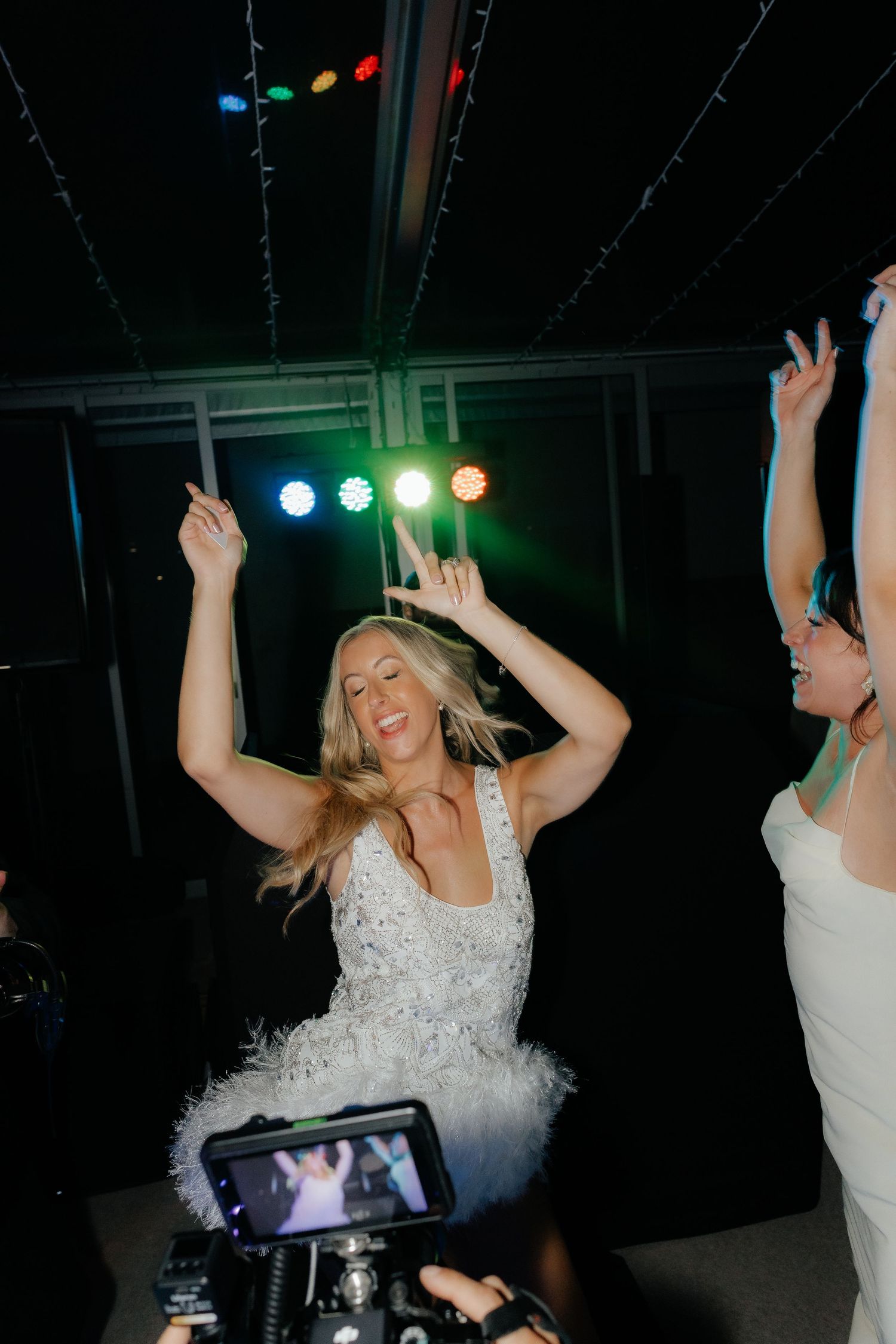 A person in a white dress dances energetically at a nightclub with colorful lights in the background.