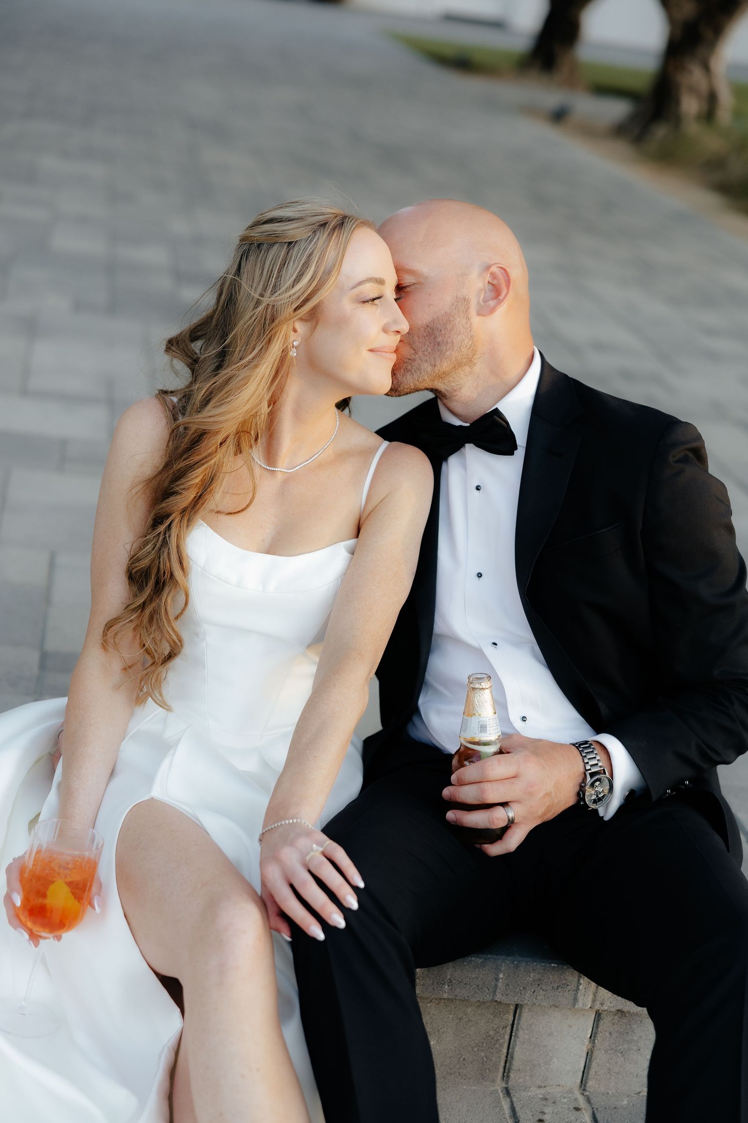 Groom kissing bride on cheek as they sit together outdoors, bride holding aperol spritz cocktail.