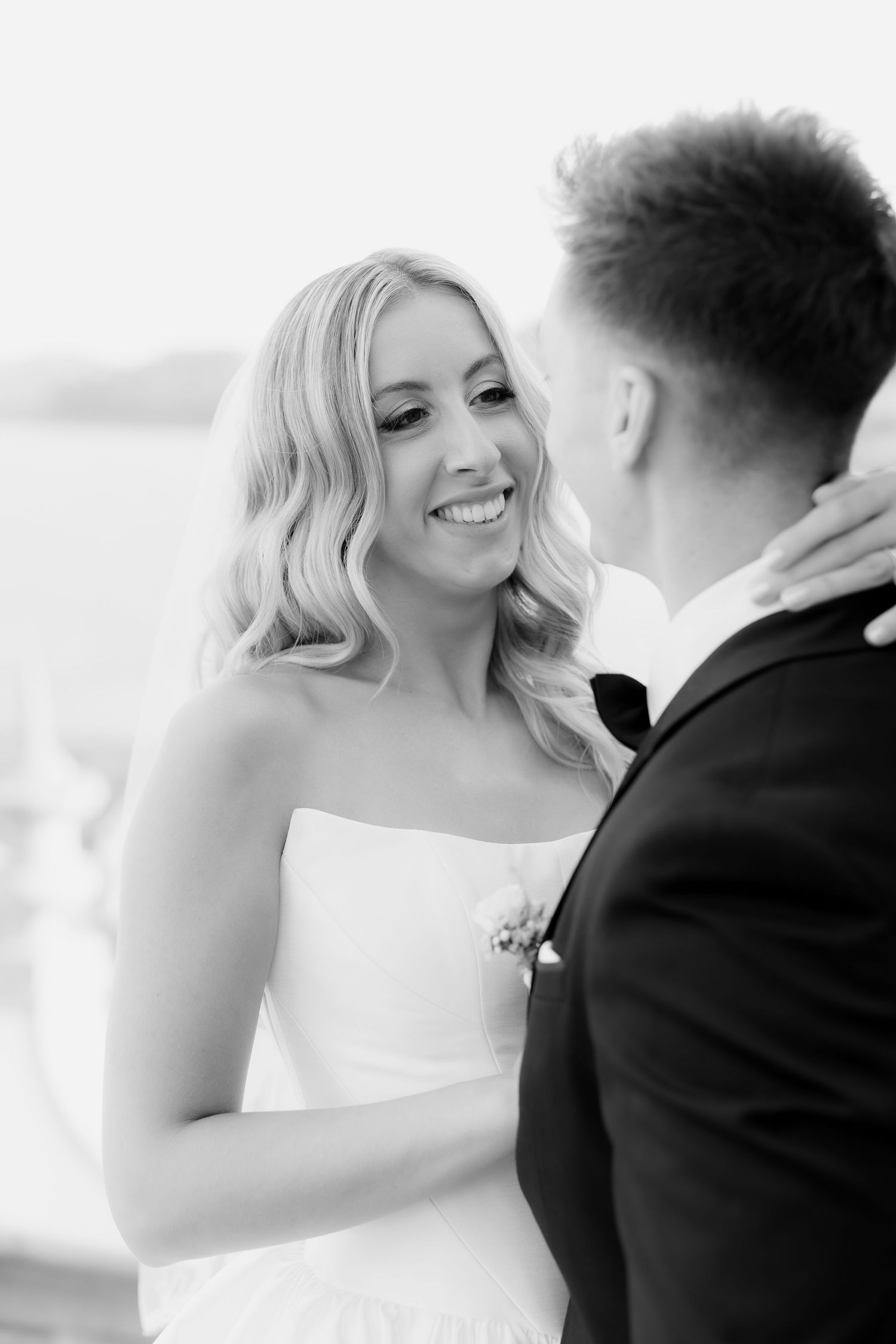 Radiant blonde bride smiles at groom in black and white close-up portrait at waterfront wedding venue.