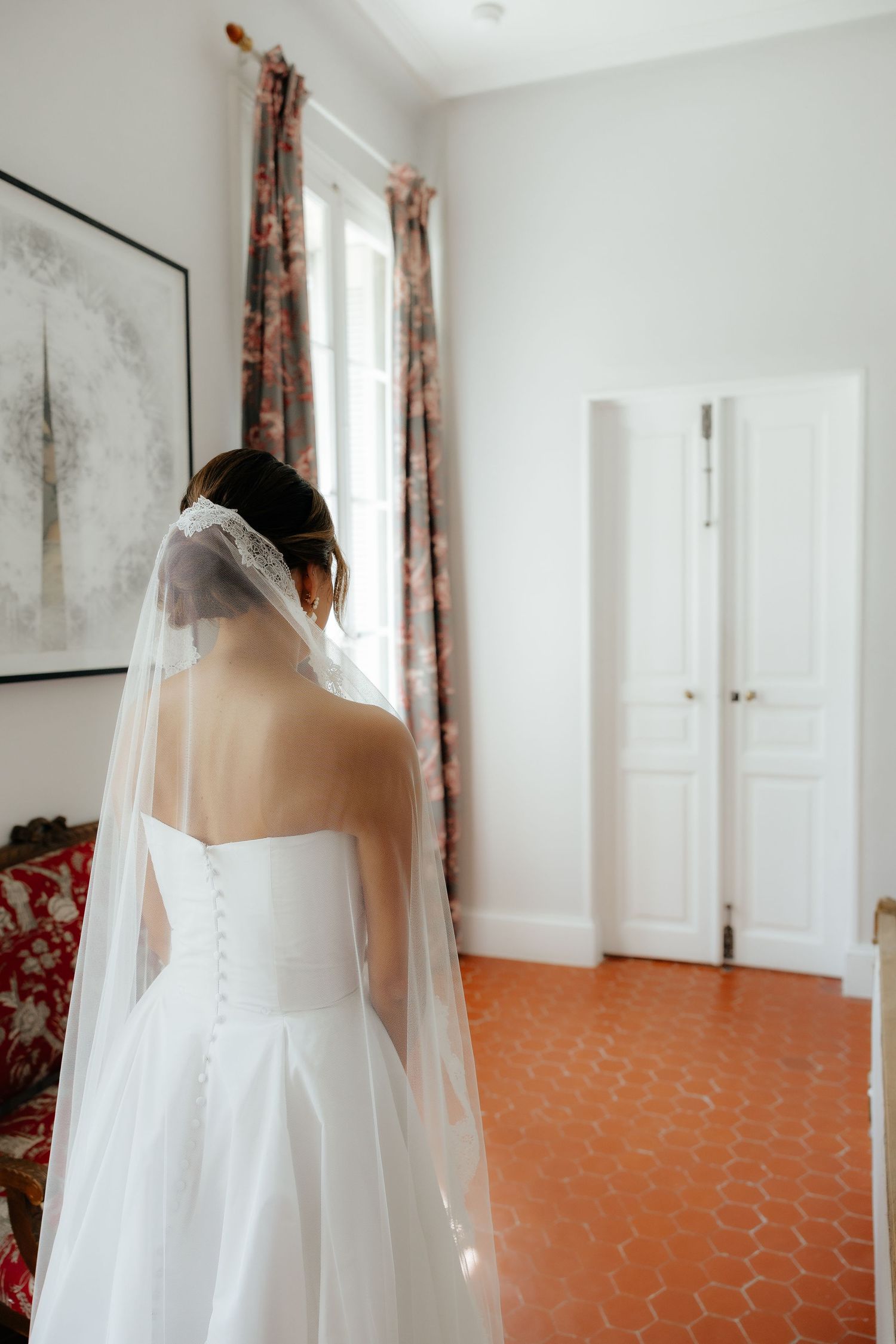 A wedding dress with delicate lace details and flowing veil shown in an elegant room with floral curtains.