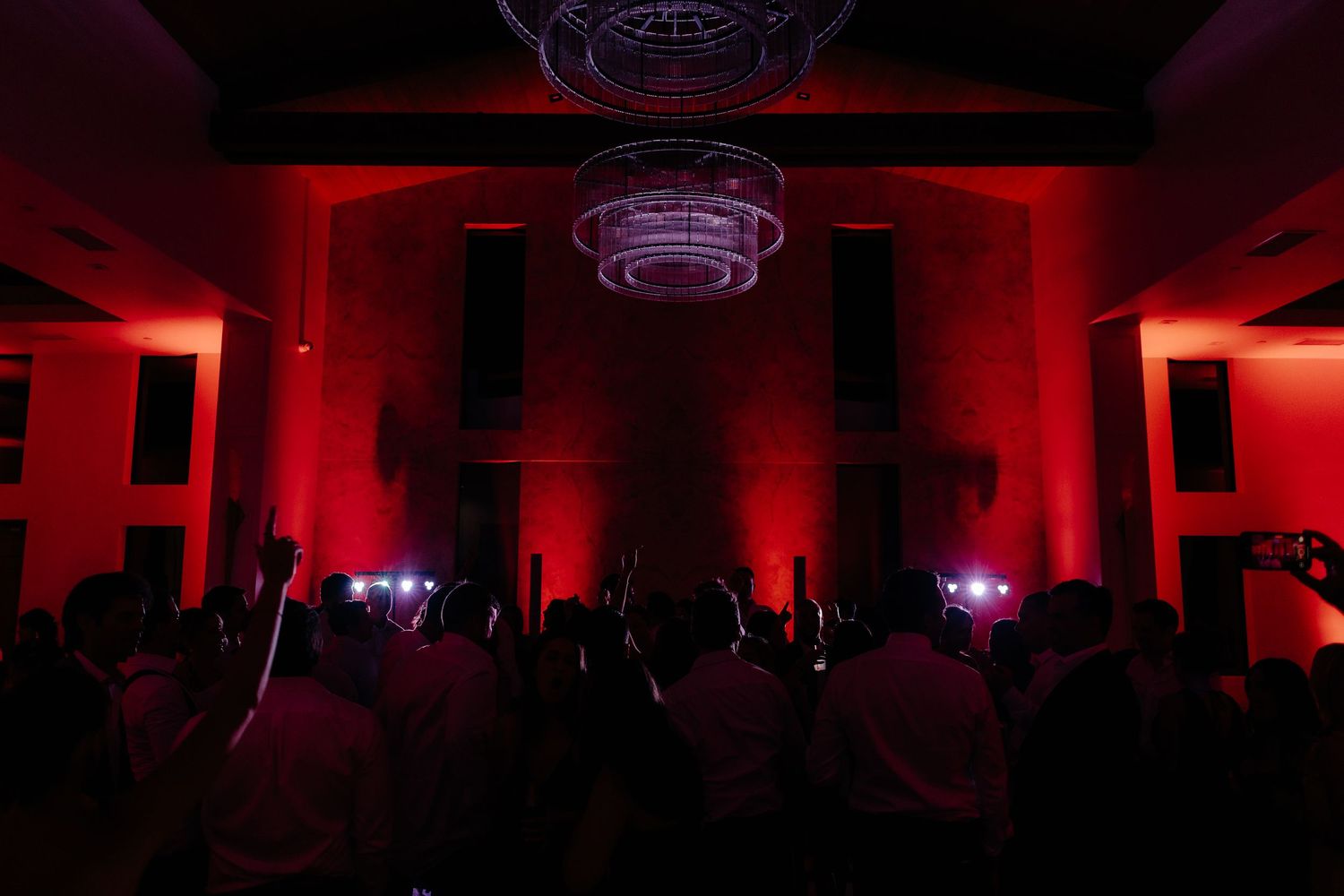 Dramatic red-lit wedding reception venue with chandelier and crowd of guests dancing with hands raised.