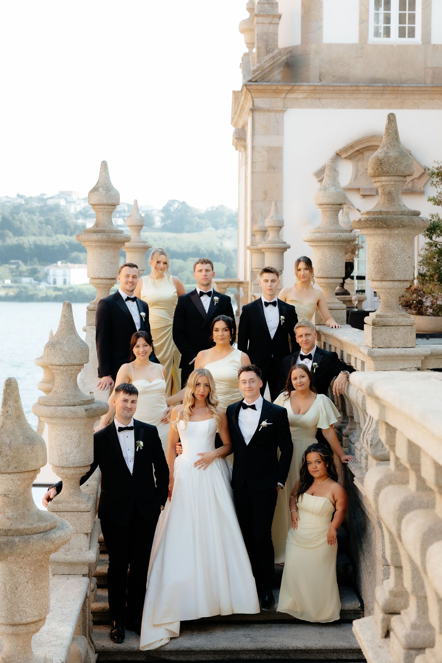 Wedding party poses on an elegant stone balcony with ornate white balusters against a scenic backdrop.