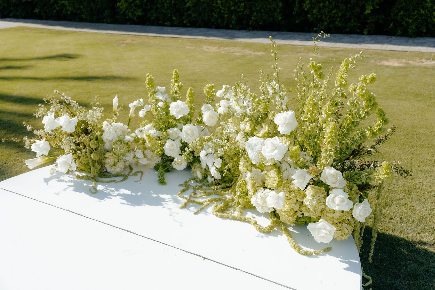 White and green floral arrangements with roses and wildflowers displayed on a white table at an outdoor wedding.