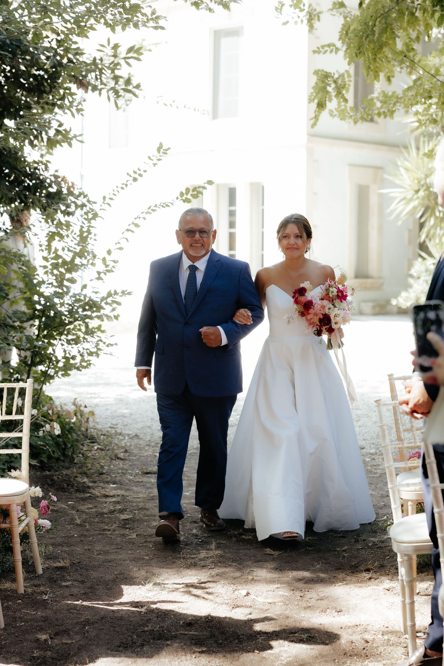 Wedding procession walking down a tree-lined path with white chairs and natural lighting.