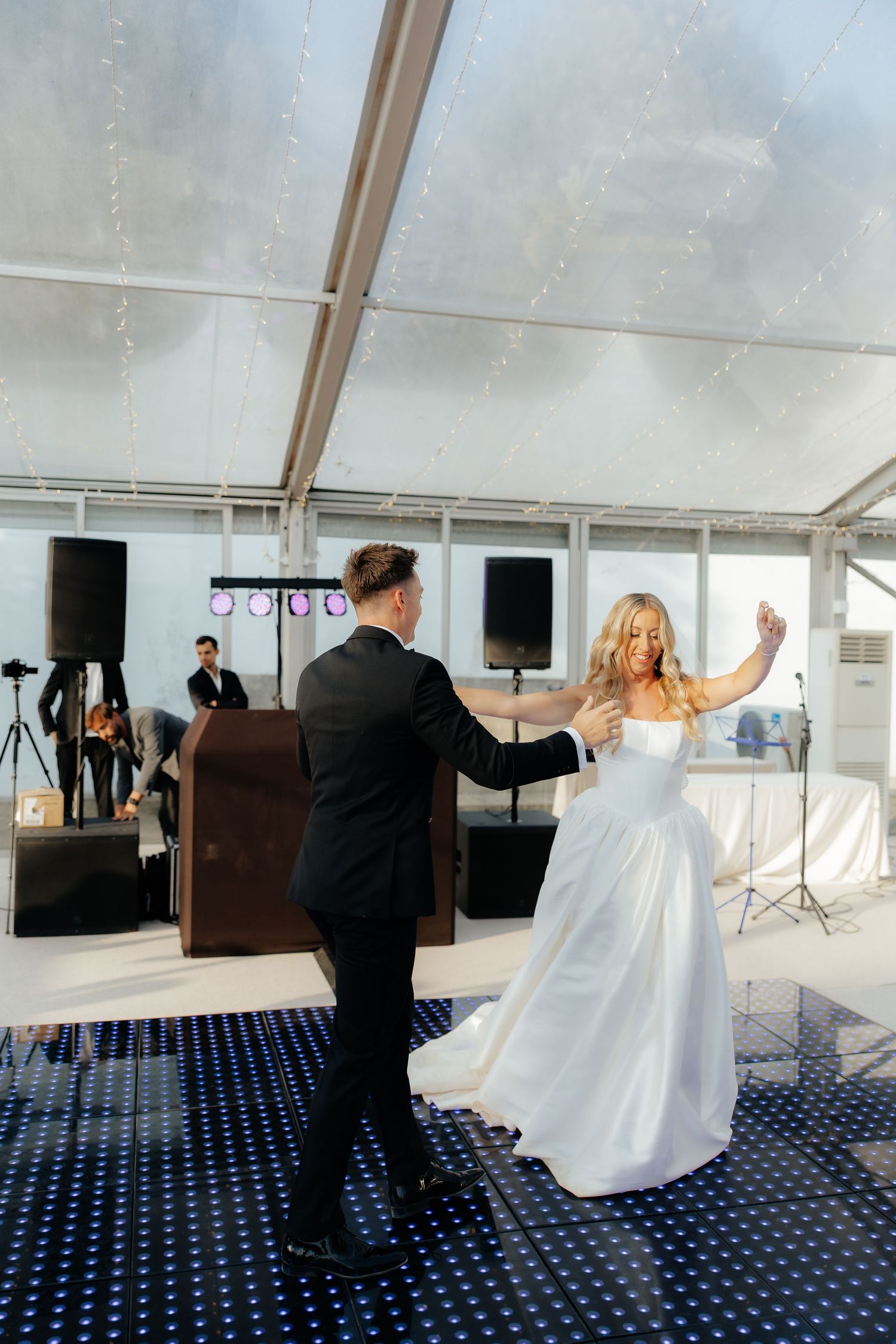 A couple in formal wedding attire dances together in a white tent with blue carpeting, captured in a sequence of photos.