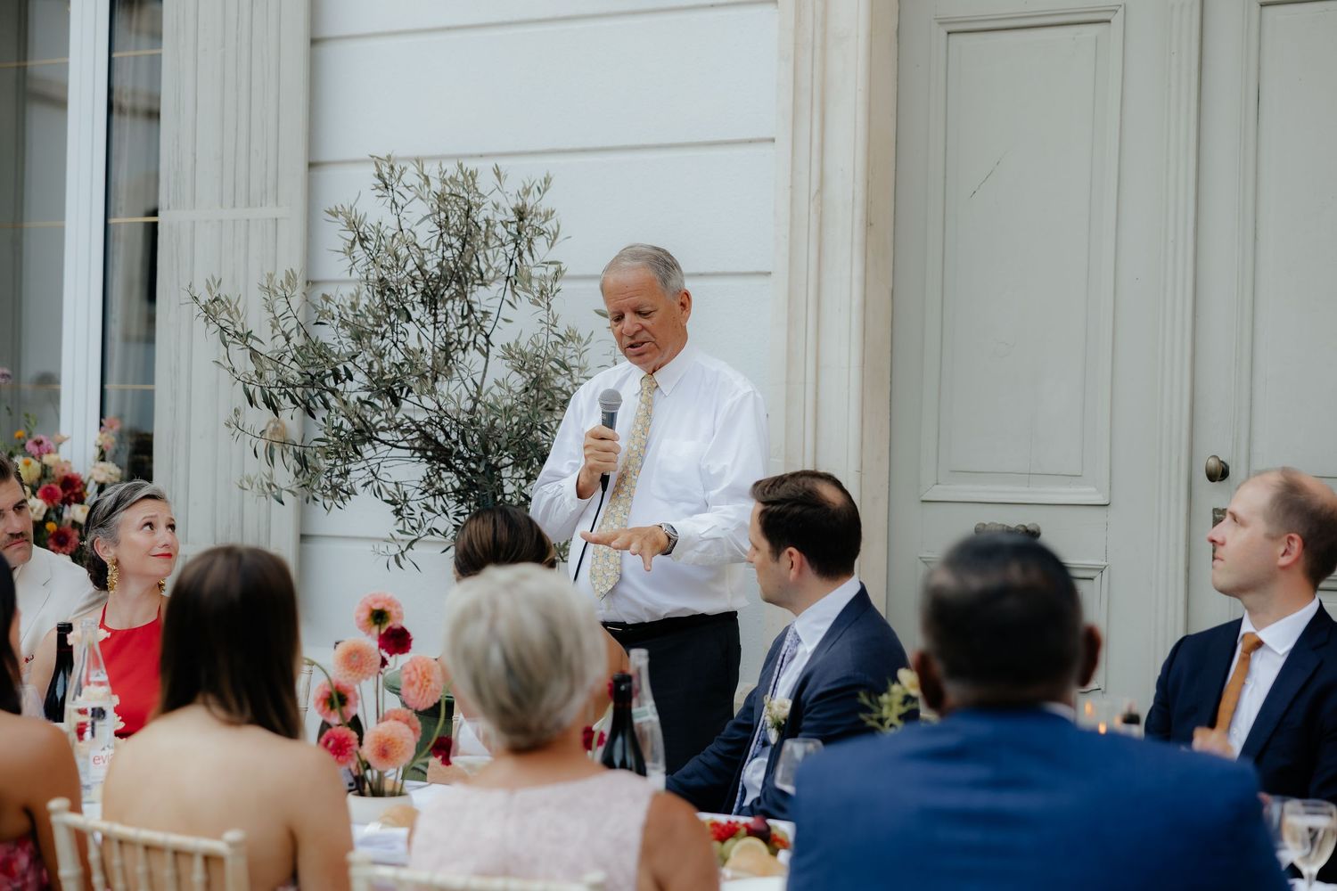 A person gives a speech during an outdoor dining event with guests seated at tables surrounded by white walls and greenery.