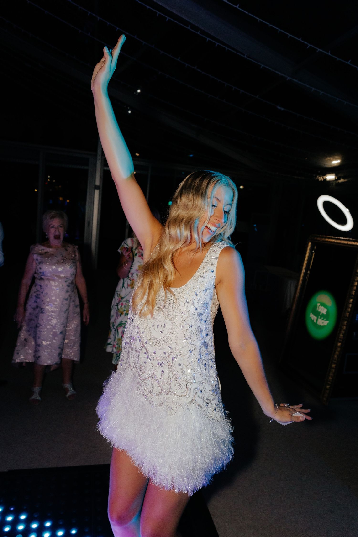 A person in a sparkly white dress dances energetically at a party with colorful lights in the background.