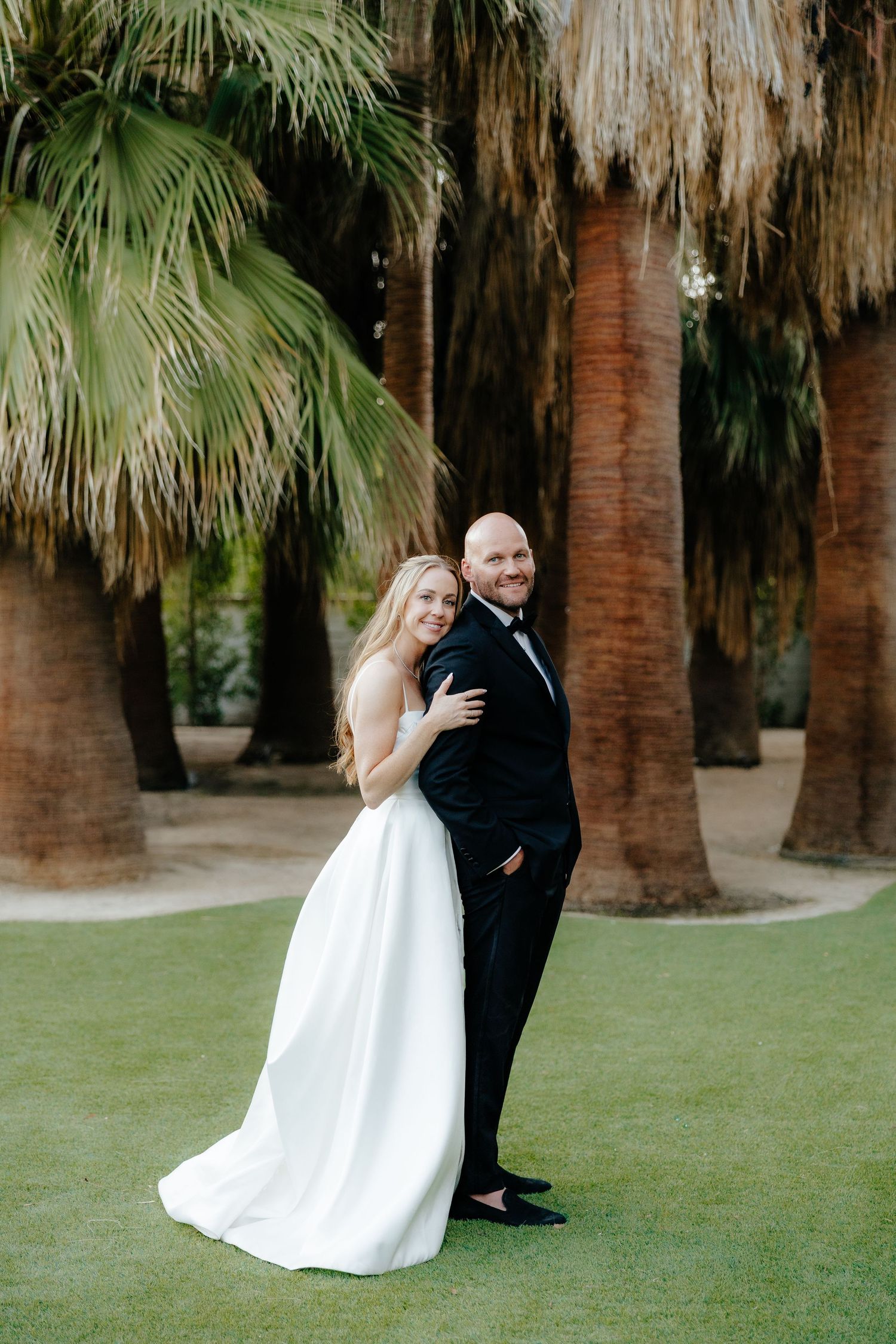 Bride and groom posing back-to-back on green lawn surrounded by tall palm trees at outdoor wedding.