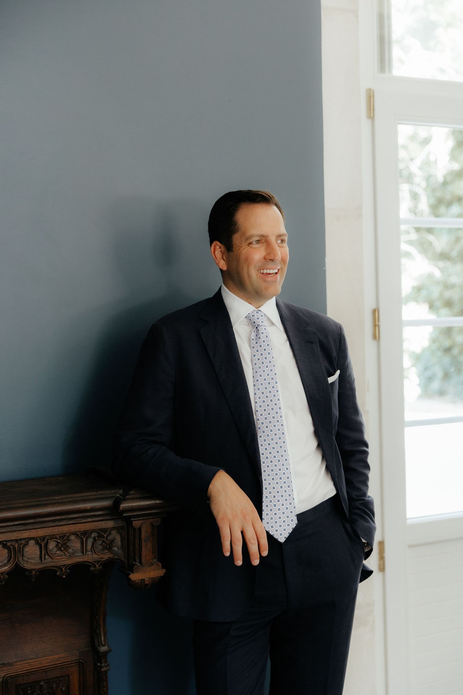 A groom in a navy suit and polka dot tie poses against a gray wall near a window.