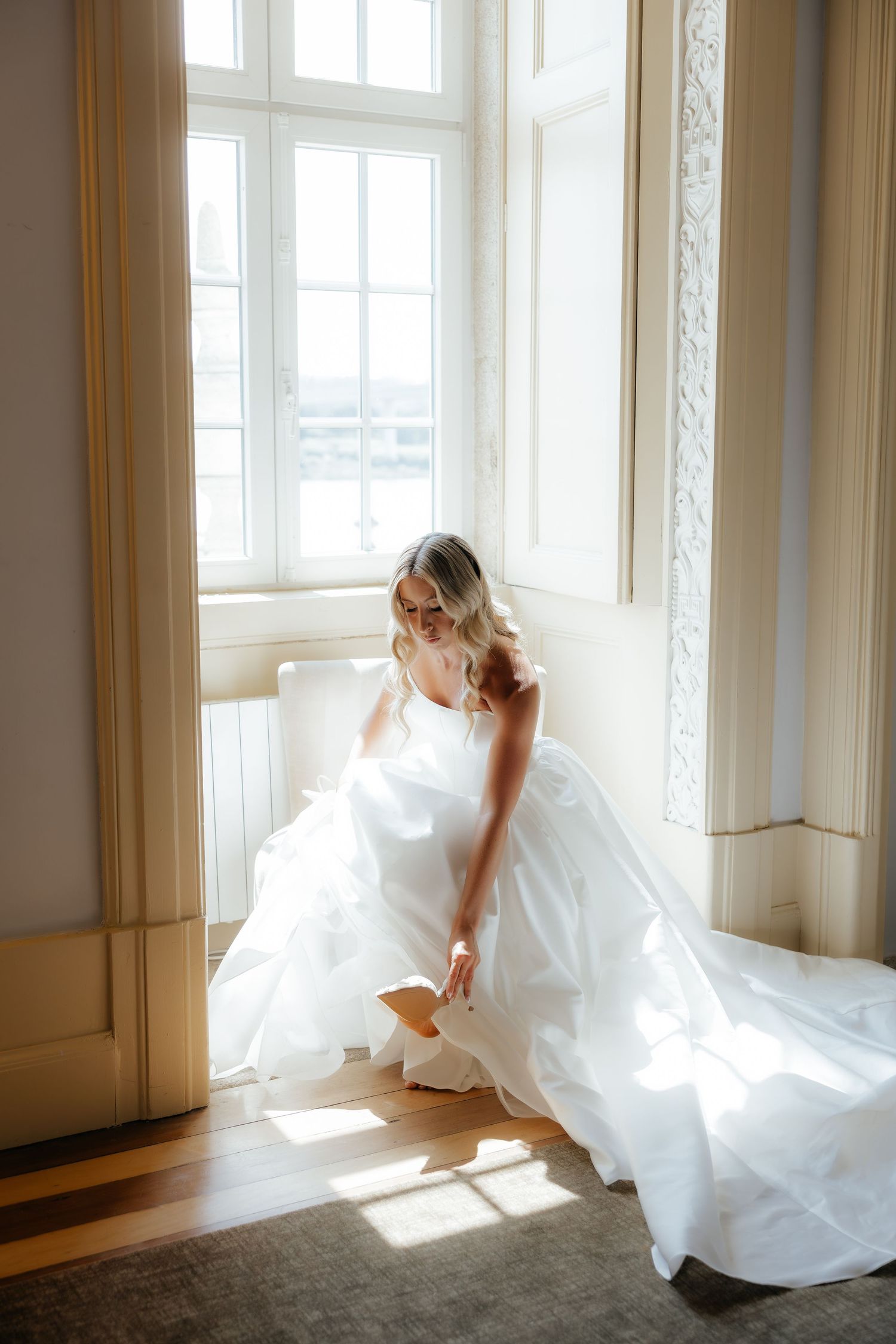 Blonde bride in white ball gown adjusting her shoe by a sunlit window in elegant venue.