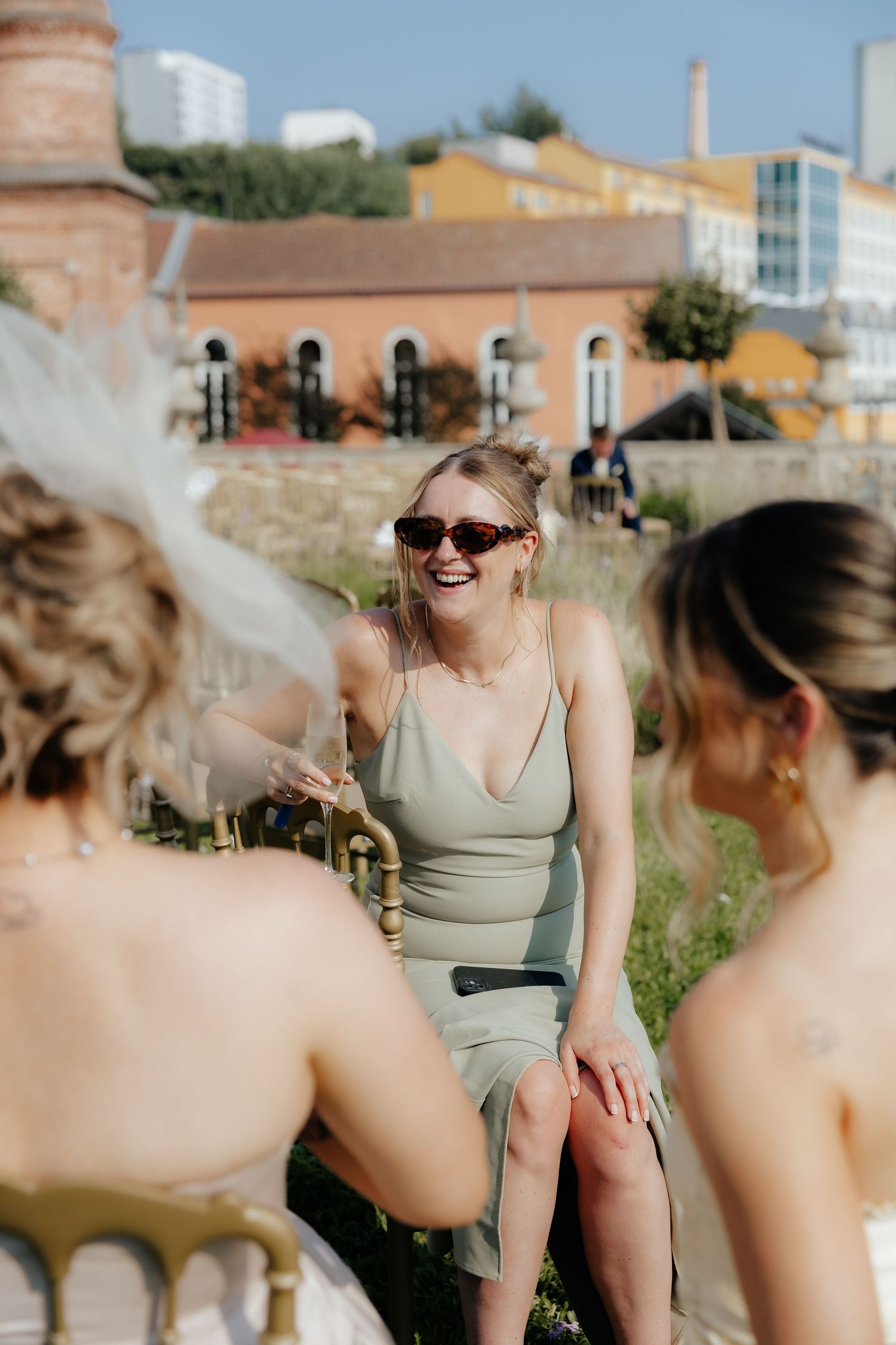 Laughing bridesmaid in sage green dress and sunglasses sits outdoors at sunny wedding celebration.