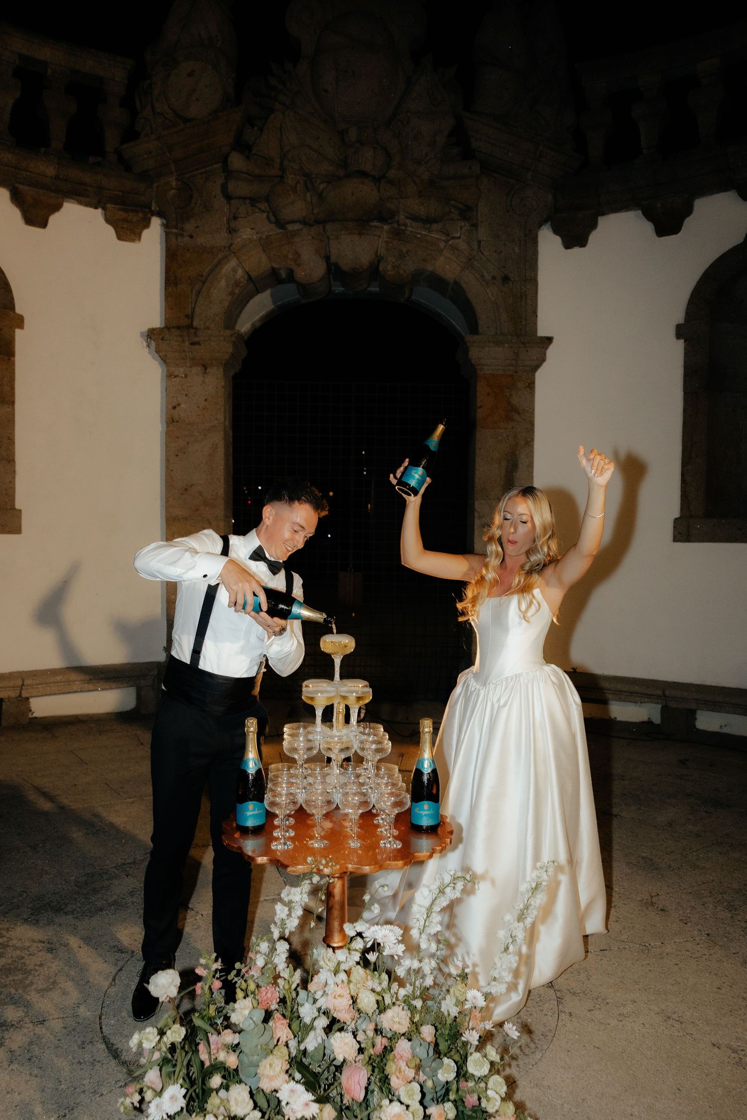 Joyful bride raises champagne bottle while groom pours at a floral-decorated champagne tower outdoors.