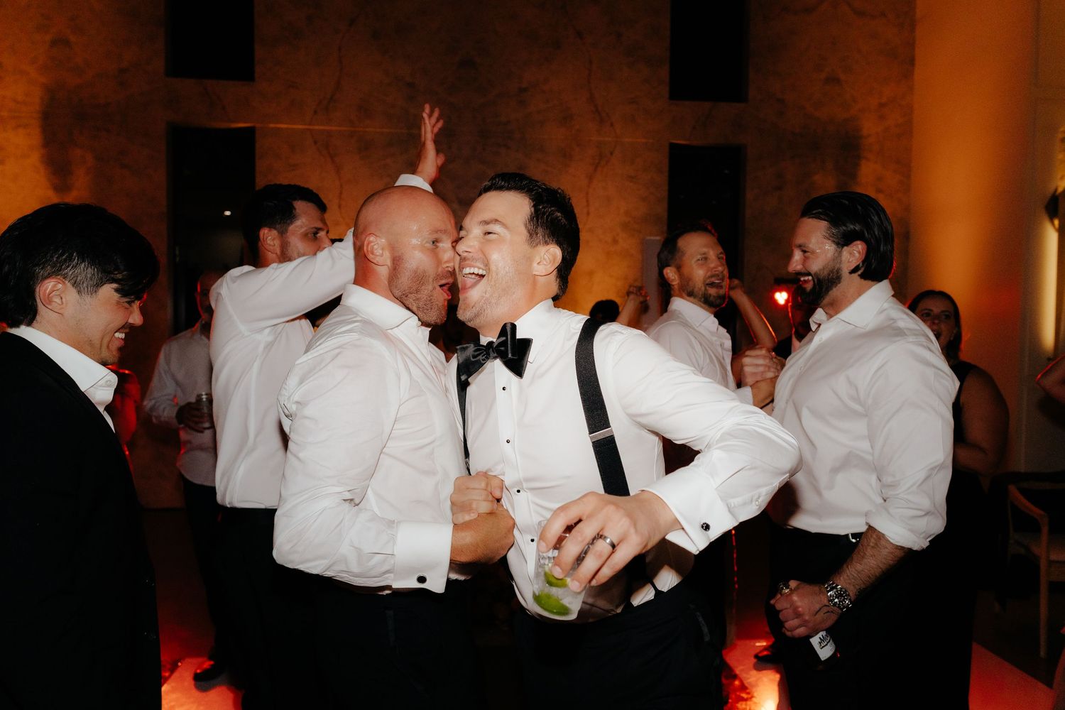 Groomsmen in white shirts and bow ties laughing and celebrating together at lively wedding reception.
