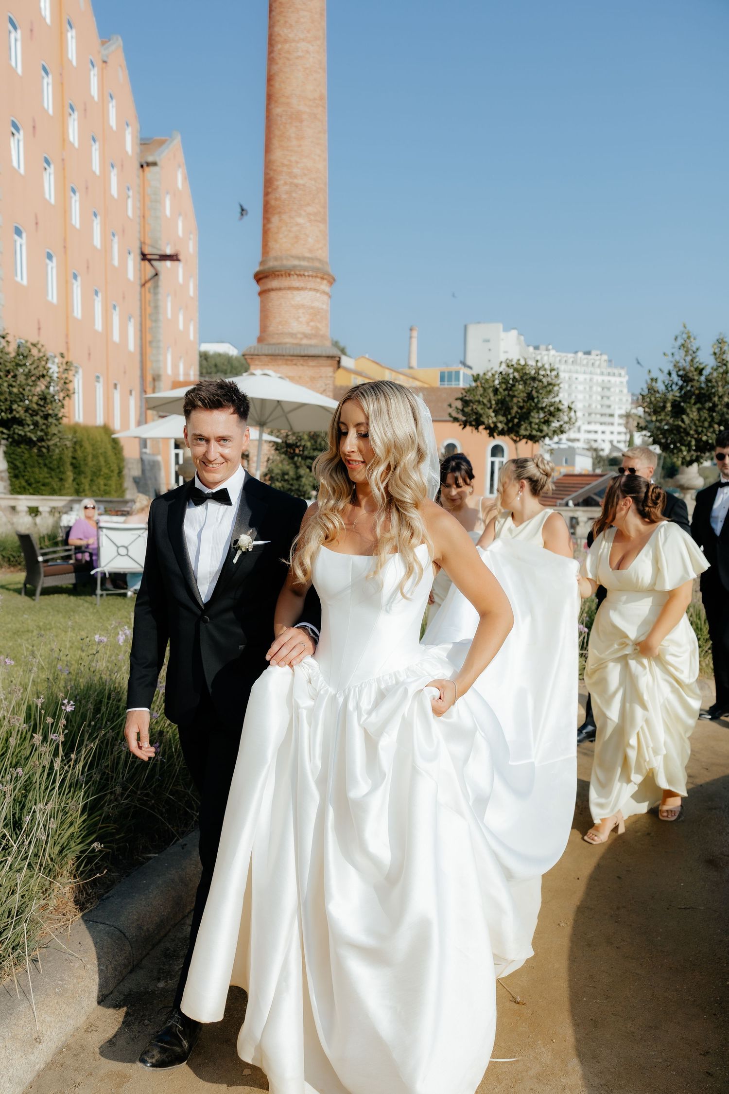 Wedding party in formal attire walks along outdoor path with bridesmaids in white dresses and groomsmen in tuxedos.