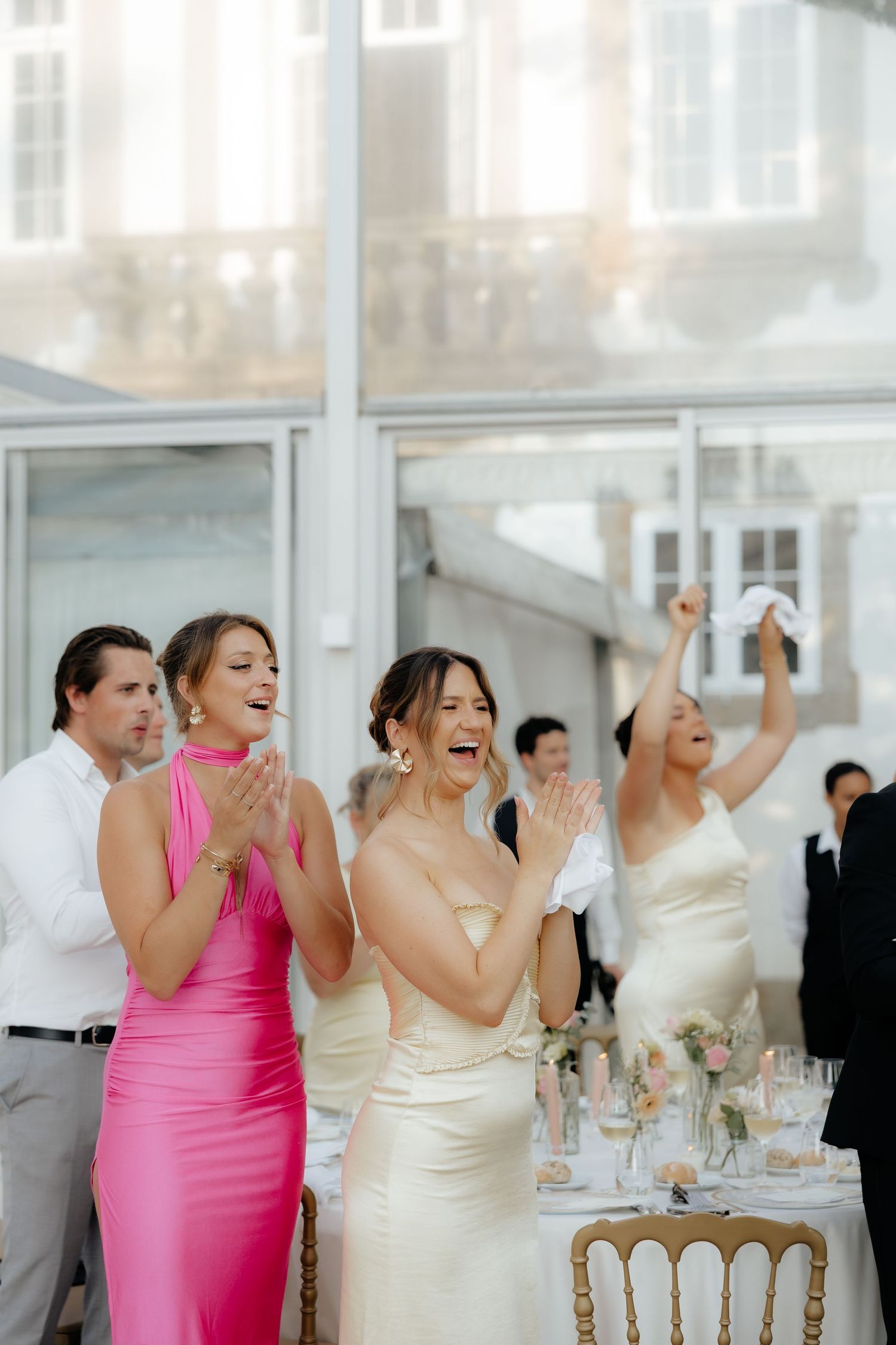 Wedding party celebrates joyfully at reception with guests raising their arms in excitement at elegant venue.