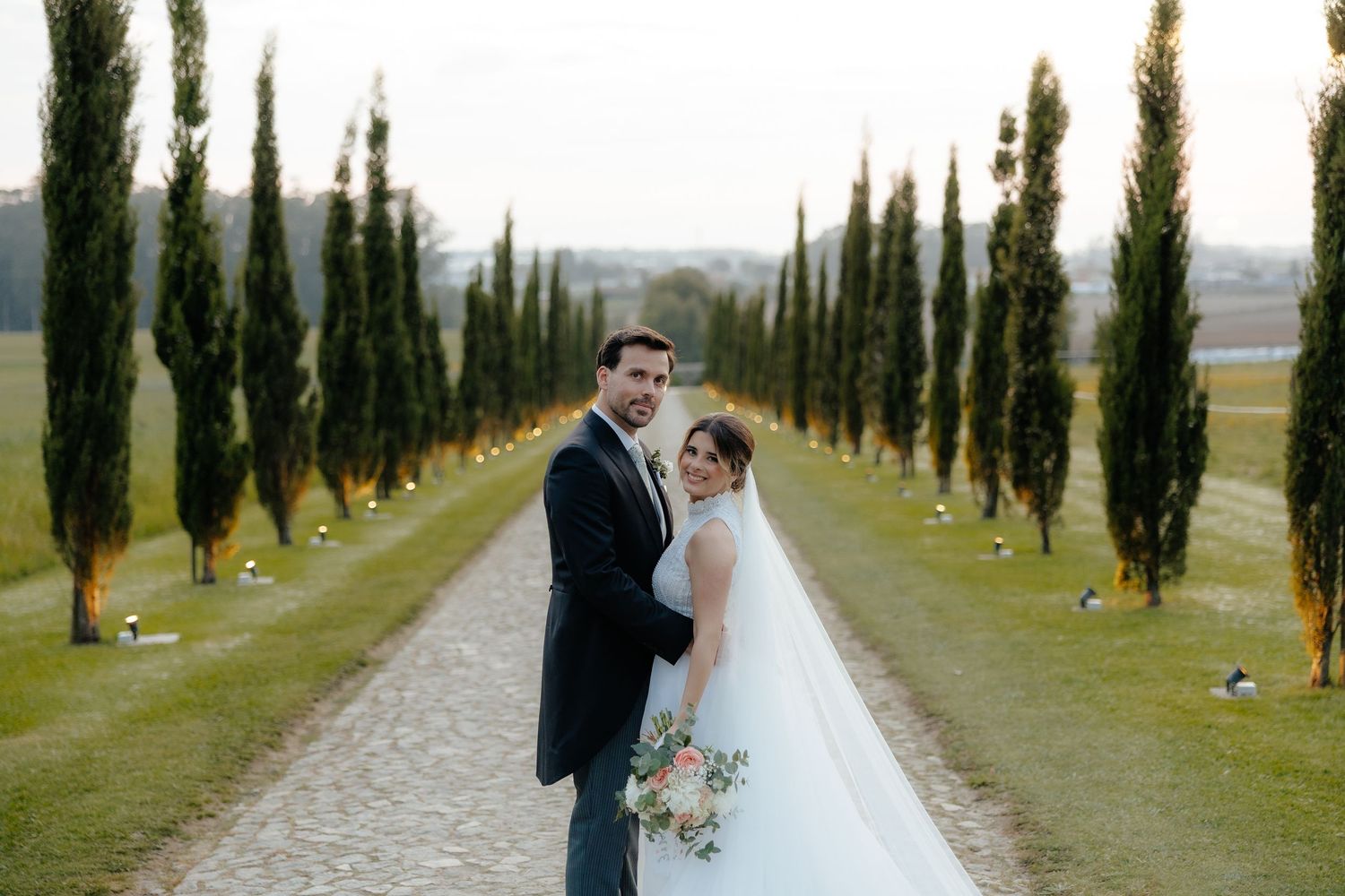 Bride and groom pose on gravel path lined with tall cypress trees at golden hour wedding venue.