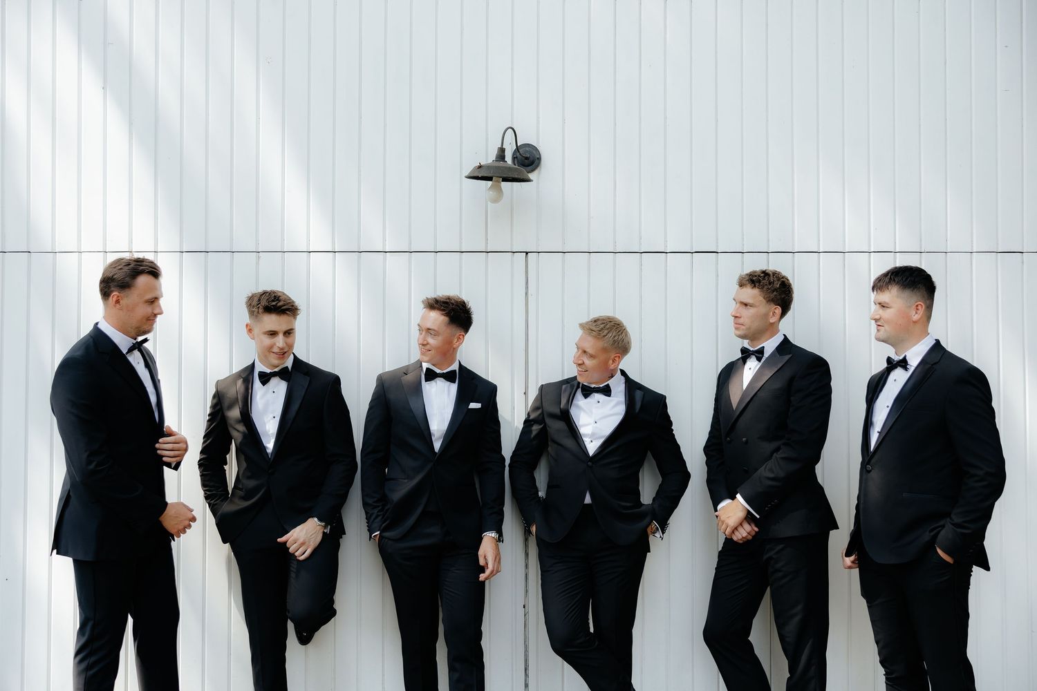 Six groomsmen in black tuxedos standing in a relaxed pose against a white barn wall.