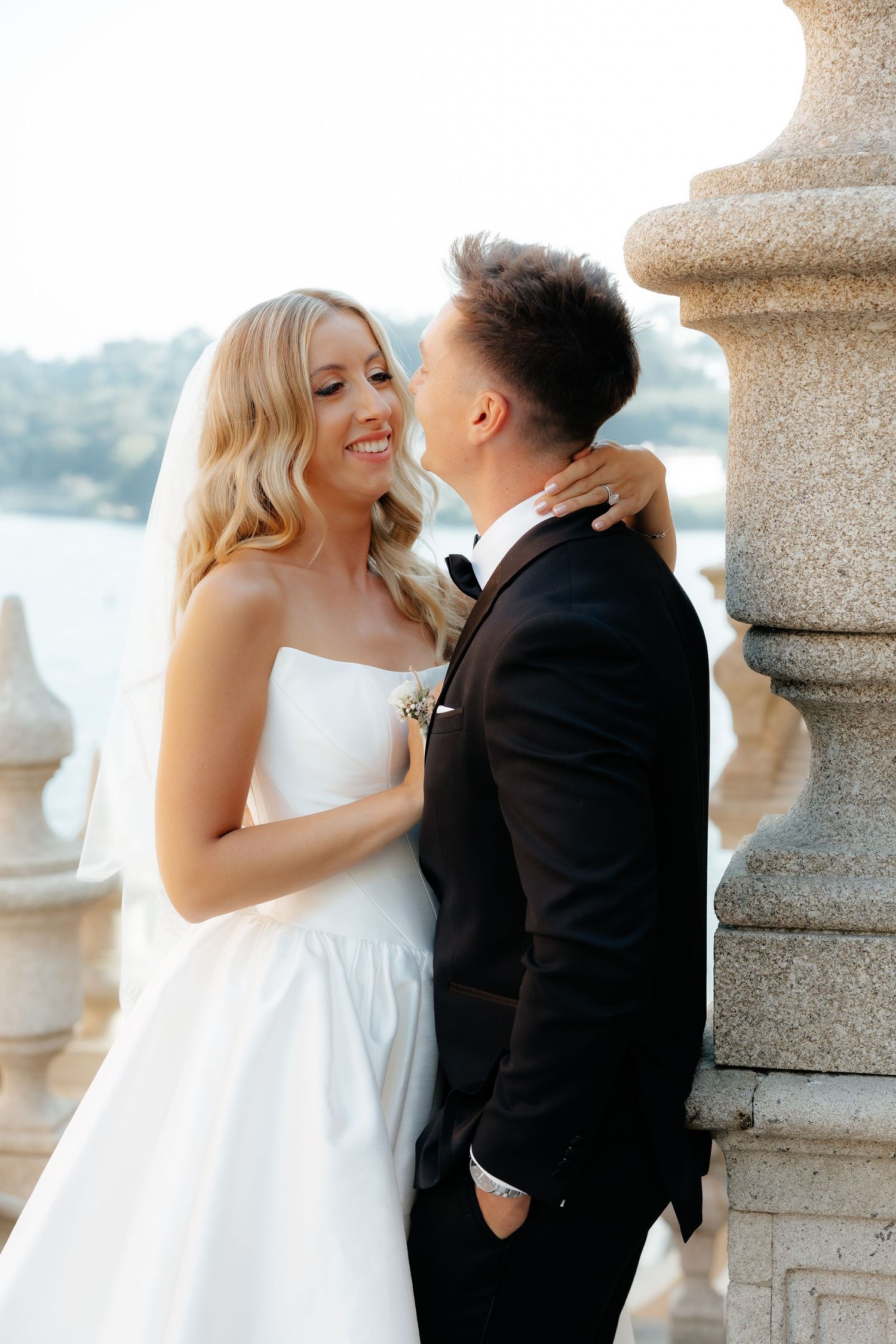 Smiling blonde bride in strapless gown embraces groom in tuxedo beside stone column at waterfront venue.