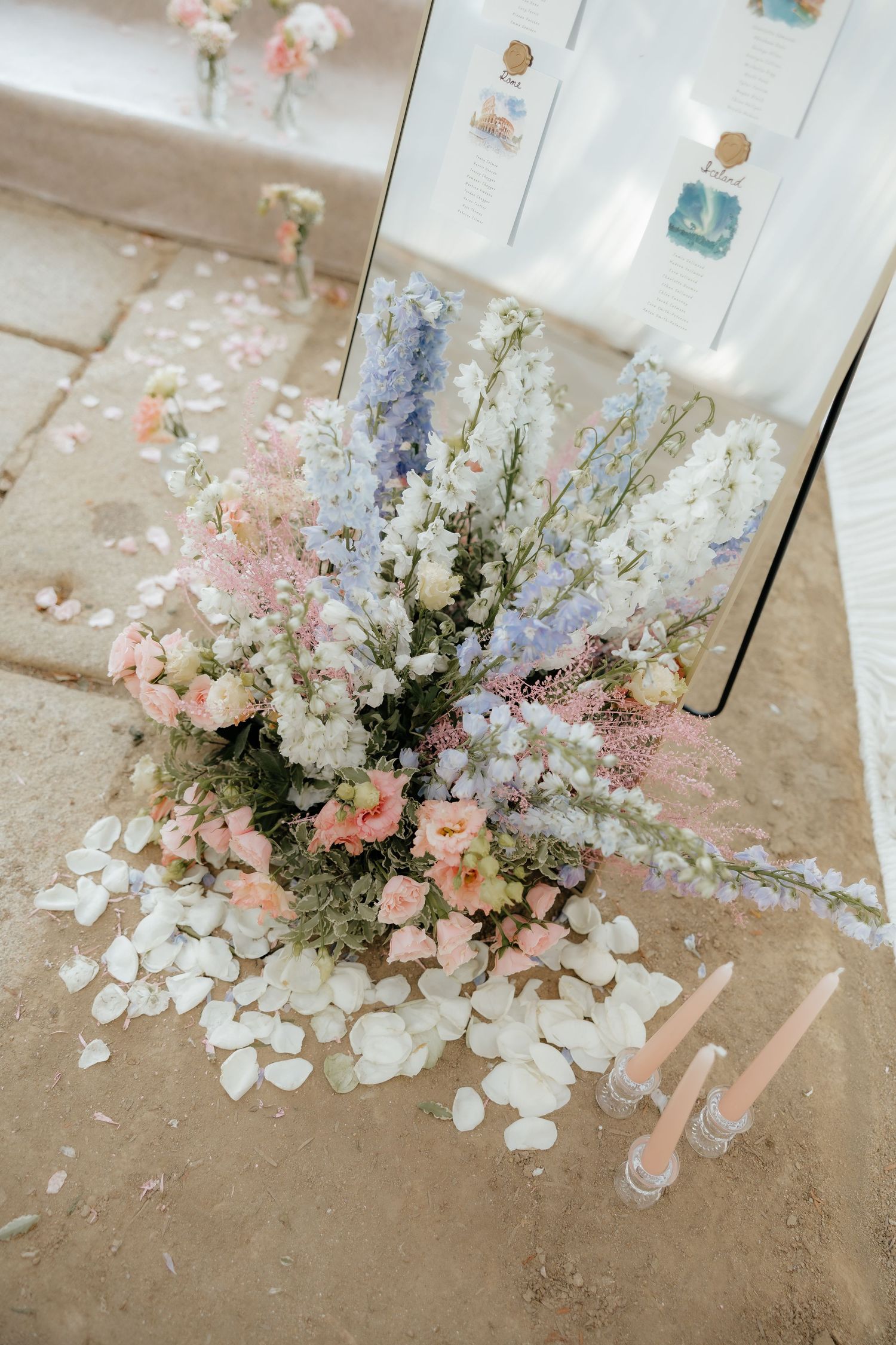 Wedding seating chart display with blue and pink floral arrangement, scattered rose petals, and pink candles.