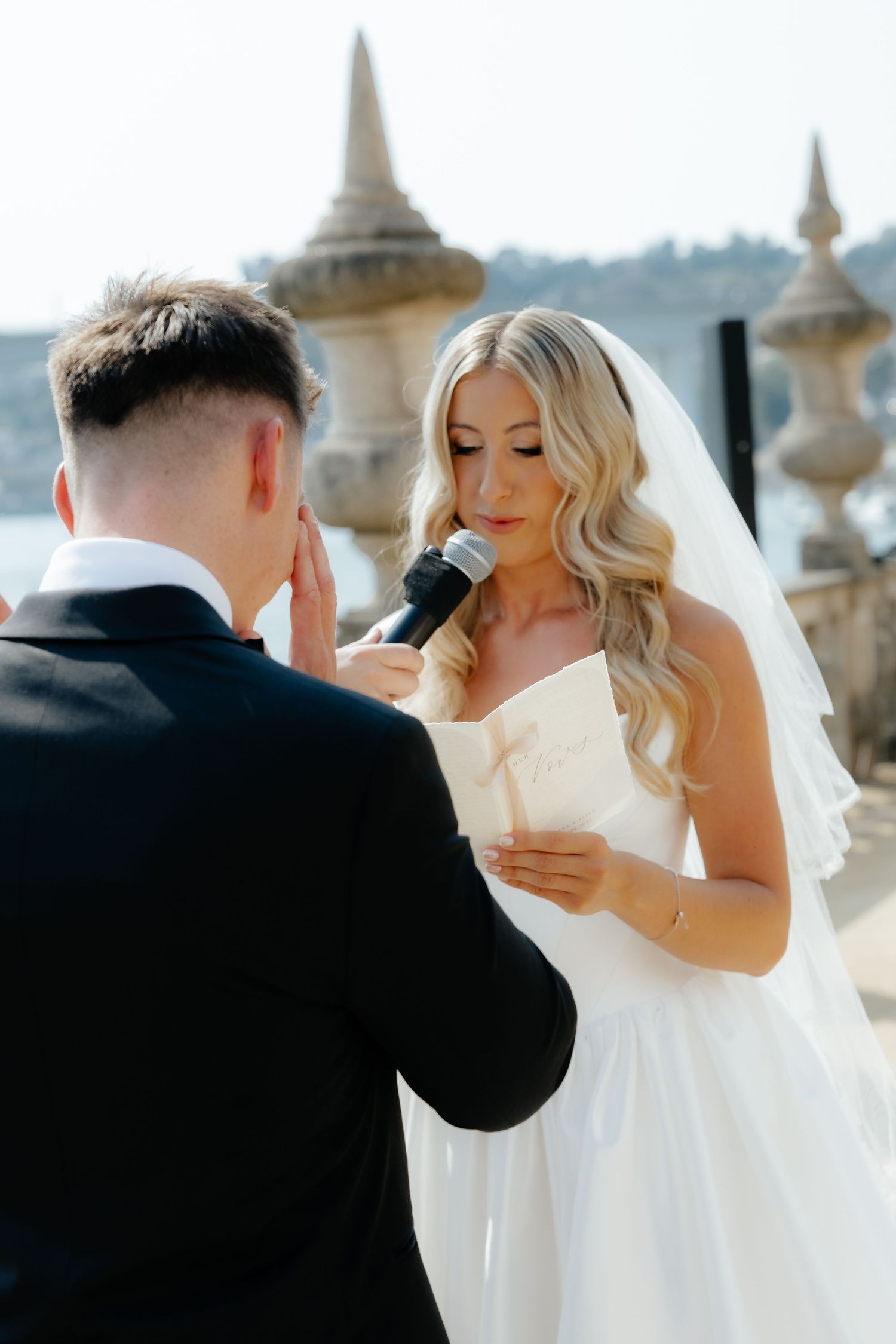Bride with wavy blonde hair and veil reads wedding vows into microphone while groom listens attentively.