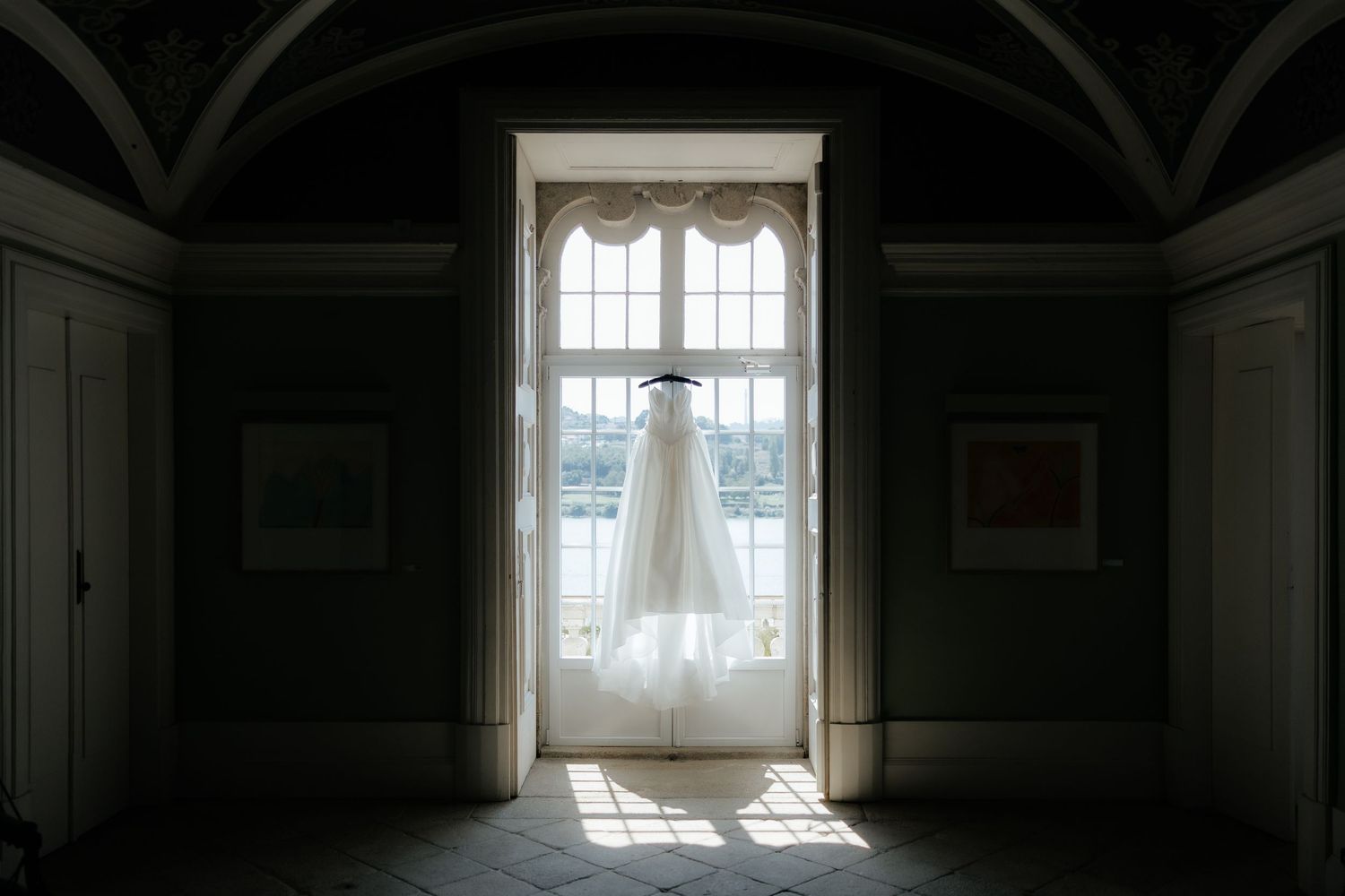 Elegant white wedding dress hanging in a grand arched window with a scenic outdoor view in the background.