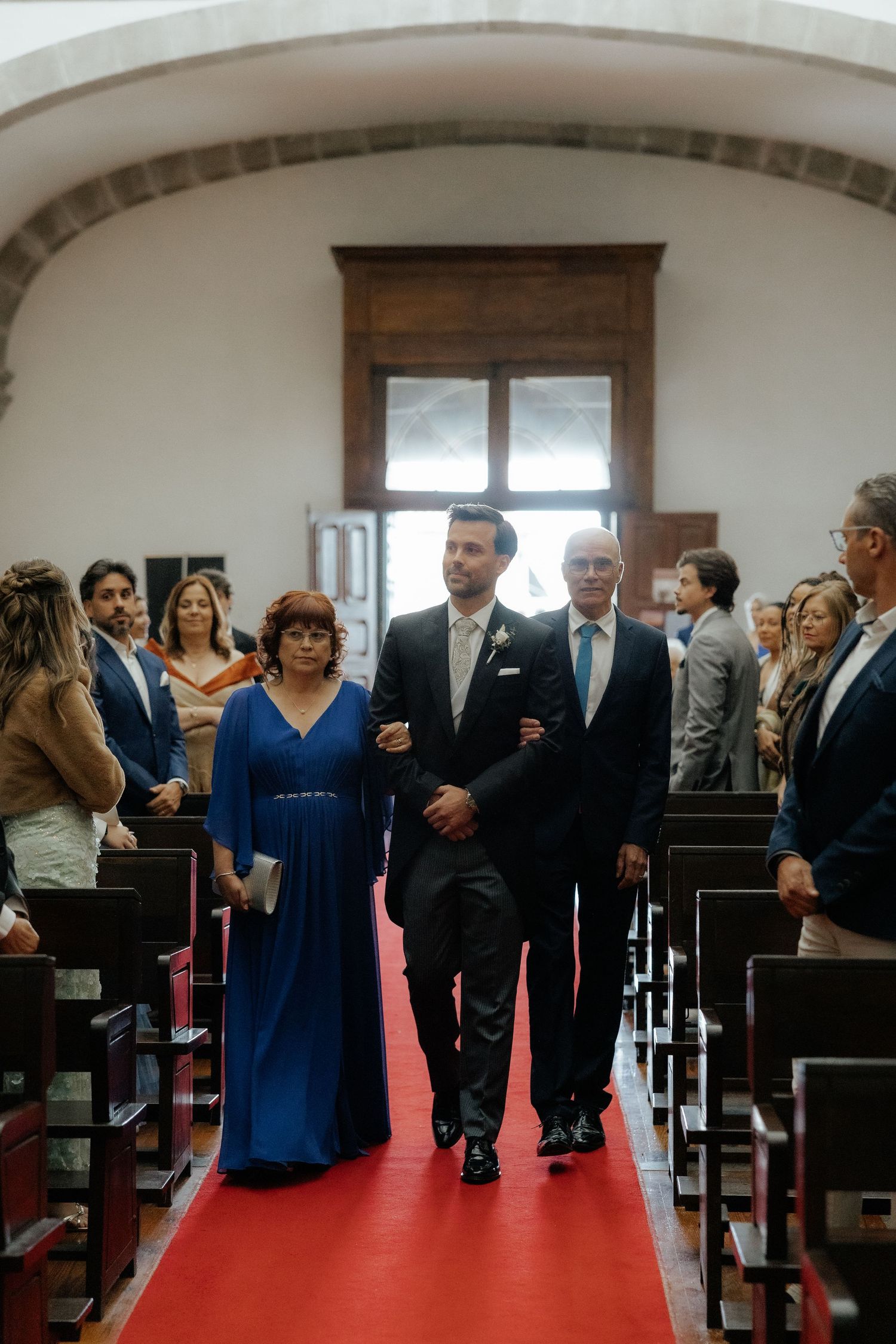 Groom walking down the red carpet aisle escorted by his parents during a church wedding ceremony.