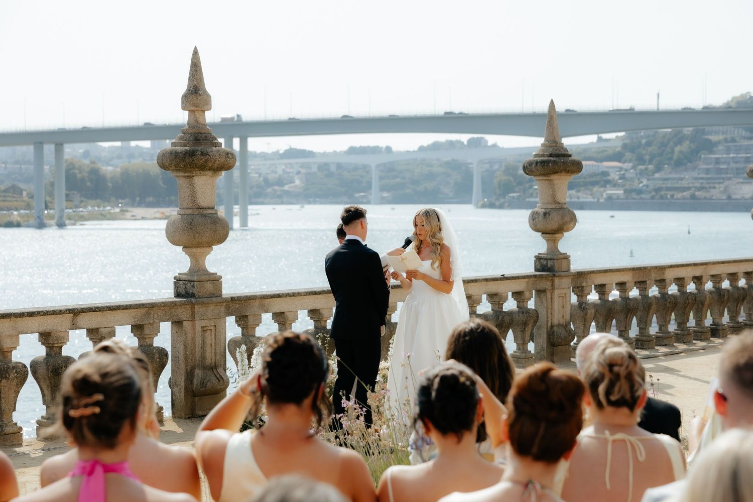 Wedding guests gather on an elegant waterfront terrace overlooking a scenic bay during a sunset ceremony.