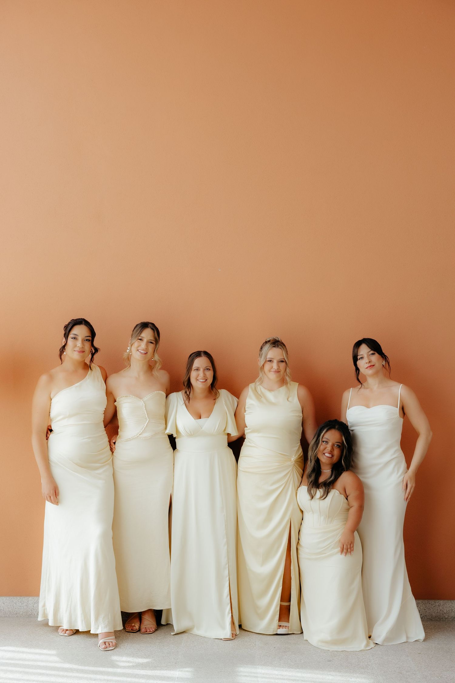Five bridesmaids in white dresses pose together against a peach-colored wall in a creative panoramic series.