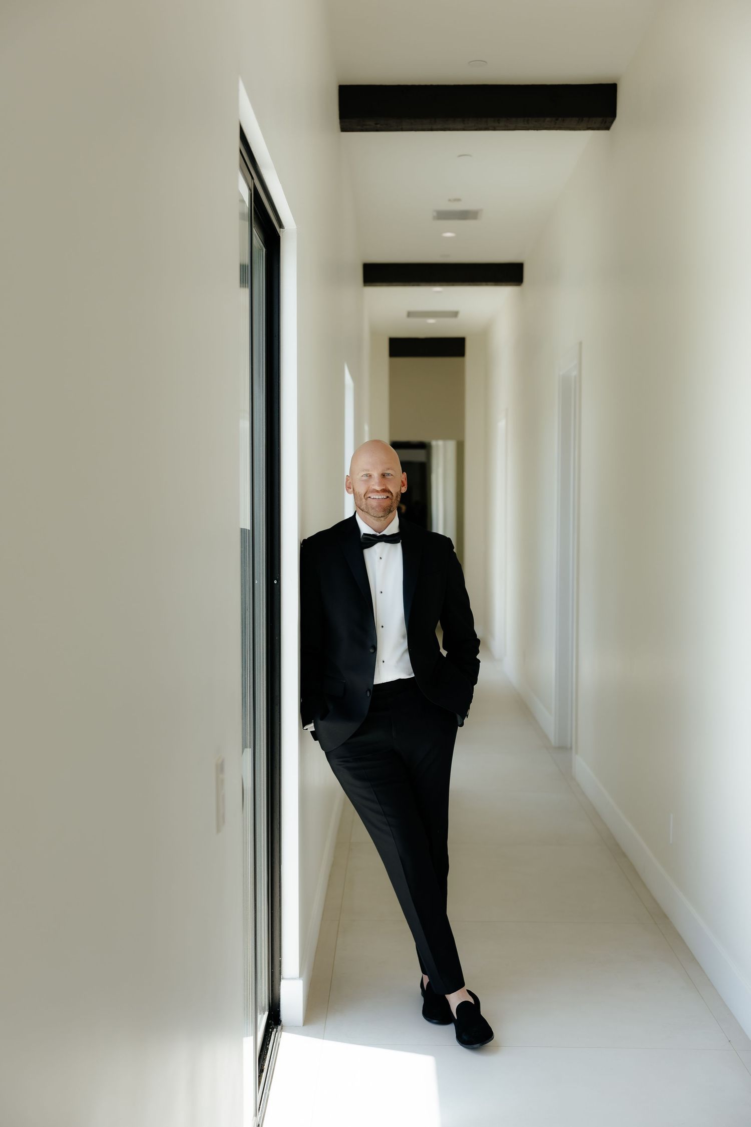 Smiling groom in black tuxedo leans against a white hallway wall in a modern bright corridor before wedding.