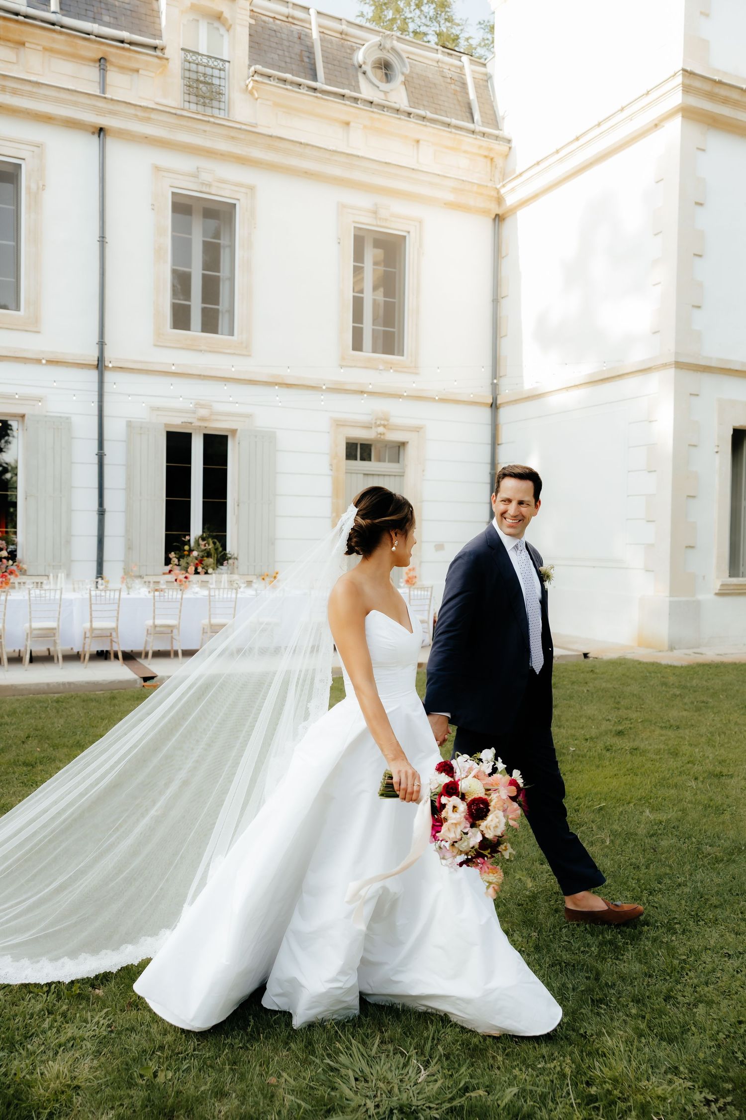A wedding couple poses in front of a grand white building with the bride's veil flowing in the breeze.