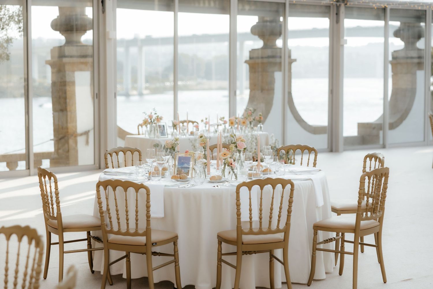 Elegant white wedding reception venue with chiavari chairs and round tables set up in a bright, window-lined room.