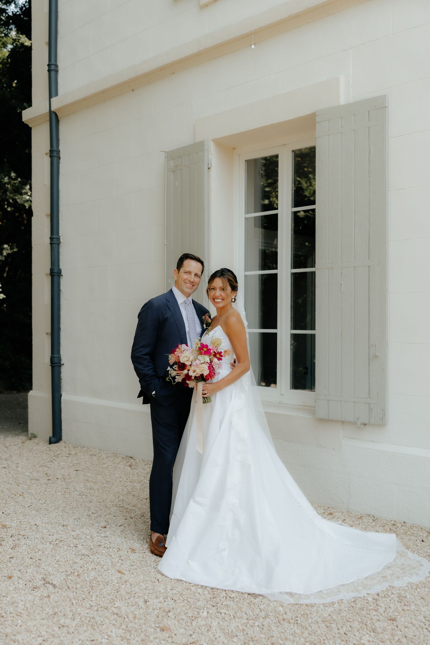 Newlyweds pose together outside white building with gray shutters on their wedding day.