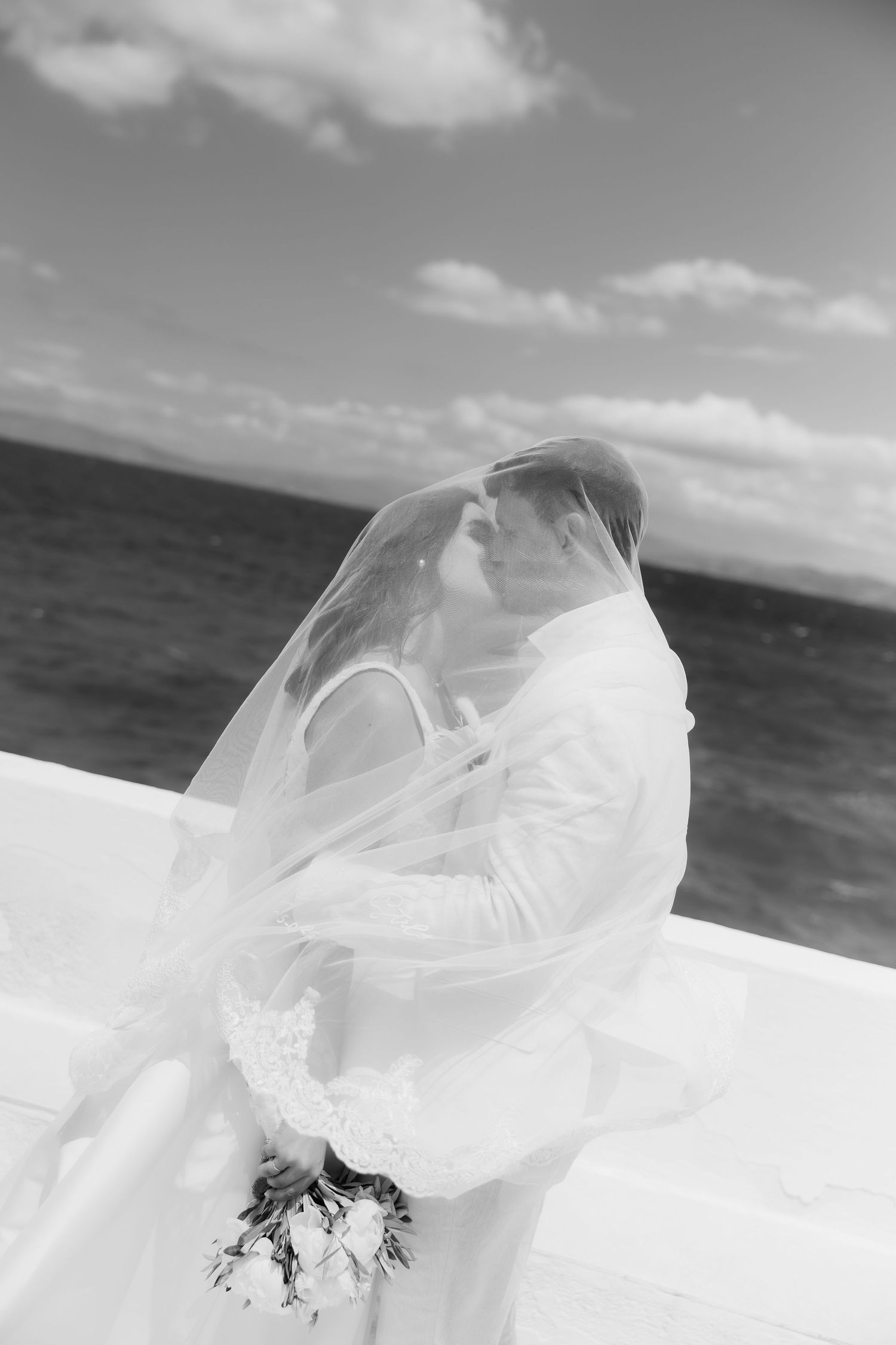 Black and white photo of a bride and groom sharing a romantic kiss under a flowing veil by the ocean.