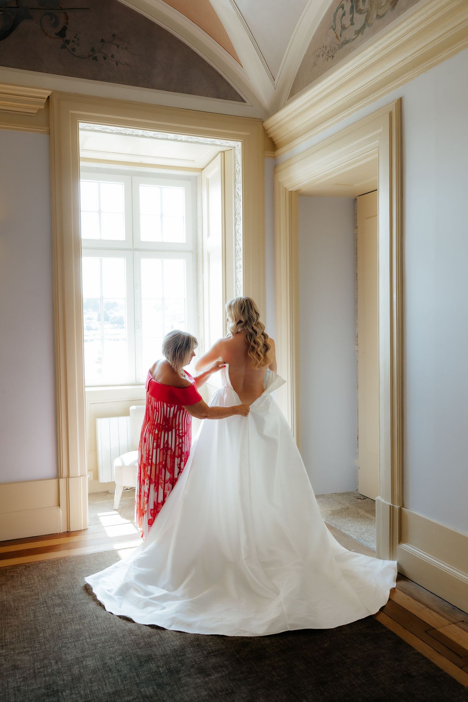 Mother in red shawl fastens buttons on bride's strapless ball gown near bright window.