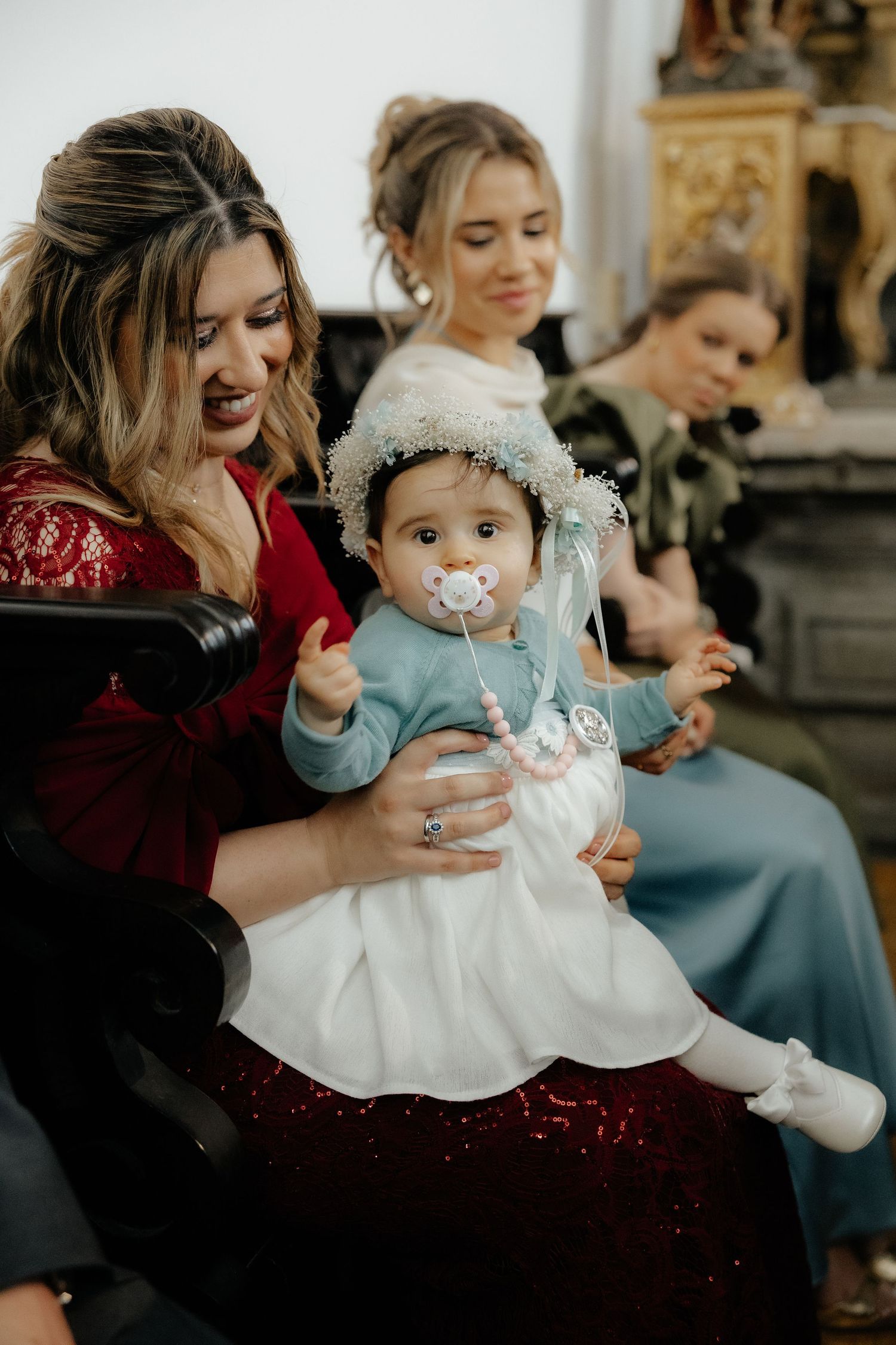 Woman in red dress holds baby in floral crown with pacifier at church wedding ceremony.