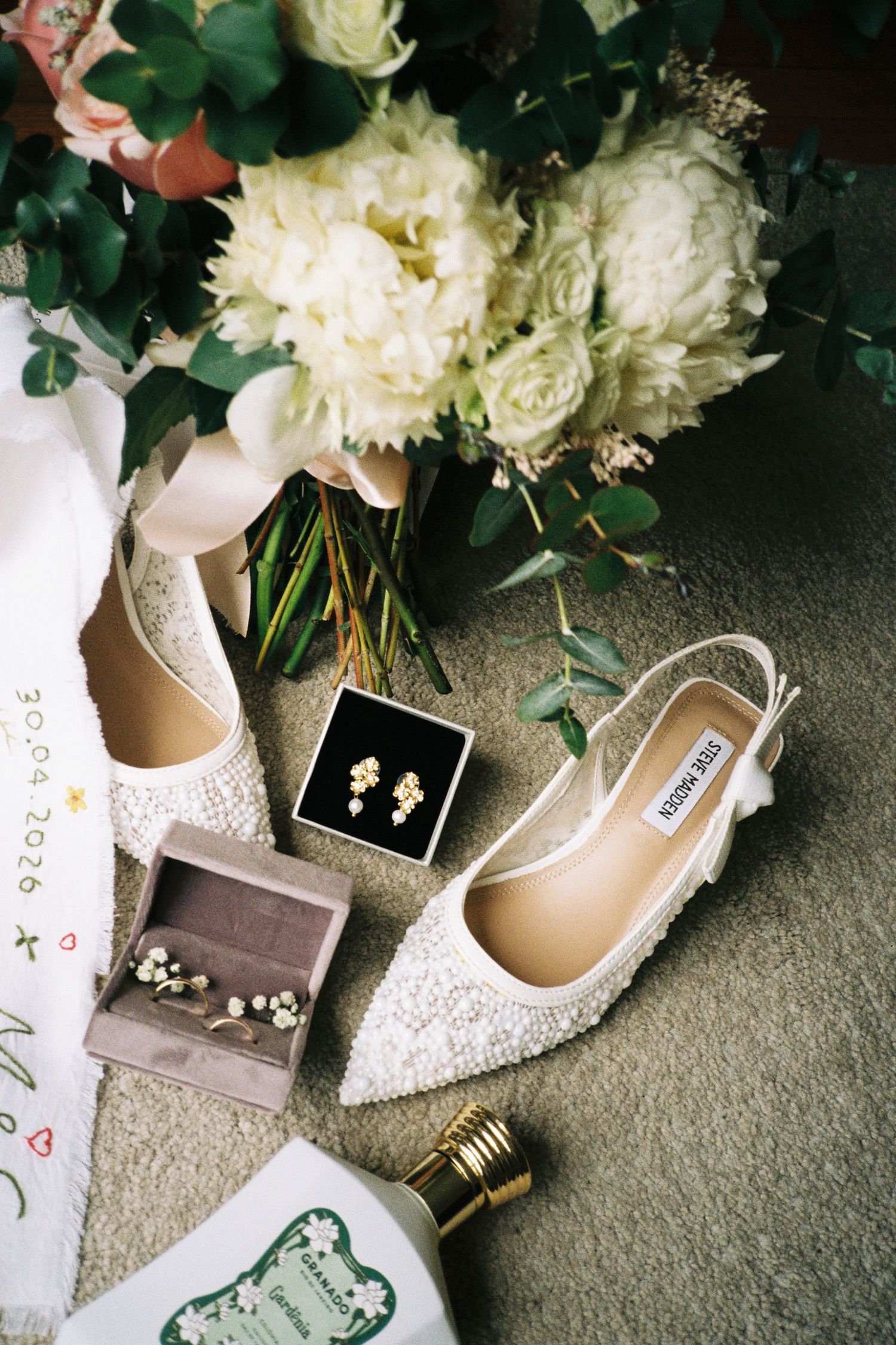 Steve Madden bridal shoes, gold earrings, white floral bouquet, and perfume flatlay display.