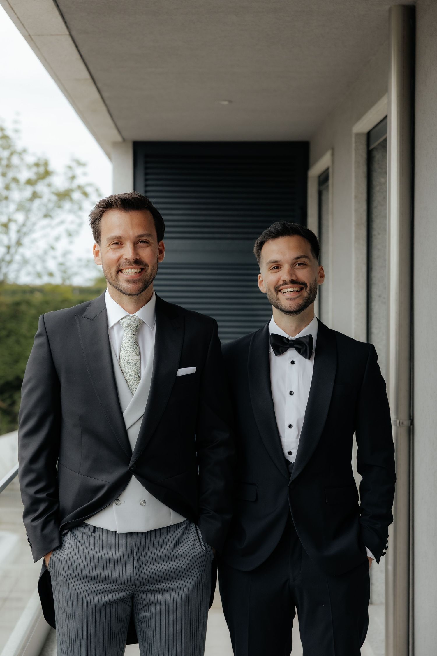 Two smiling men in formal suits pose together on a balcony outdoors.