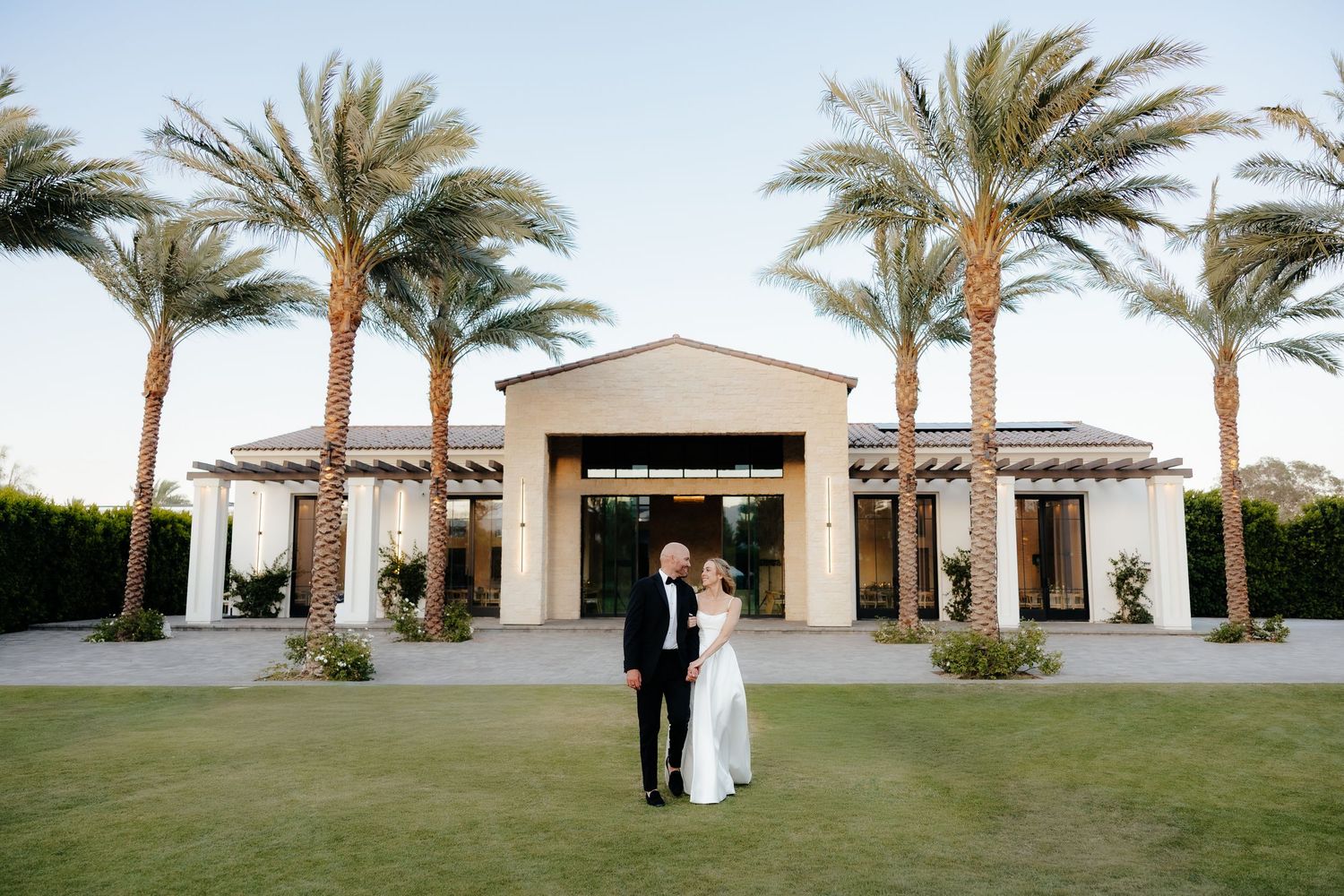 Bride and groom standing on green lawn in front of elegant palm tree-lined wedding venue at dusk.