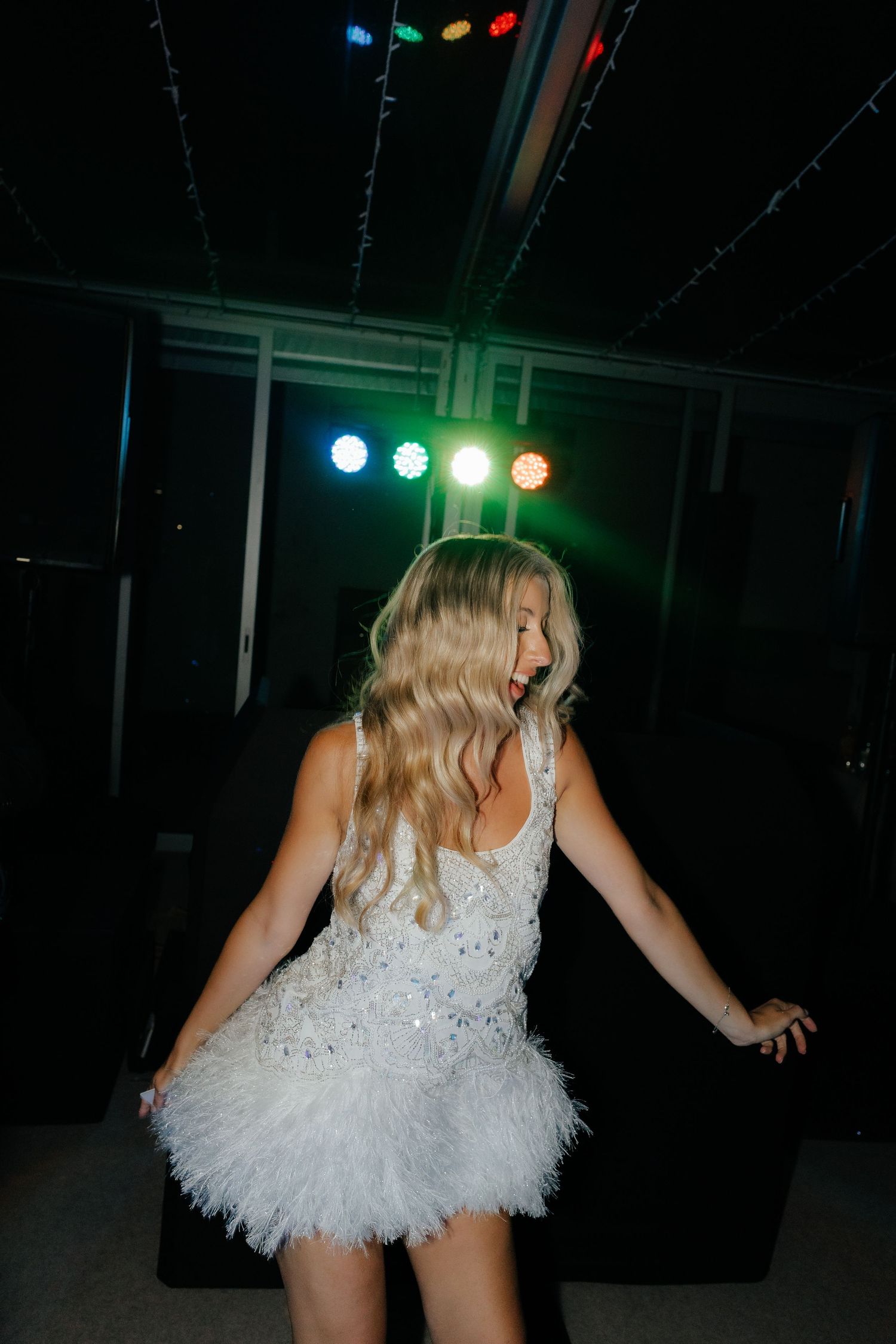 A dancer in a white feathered dress performs under colorful stage lights in a dark venue.